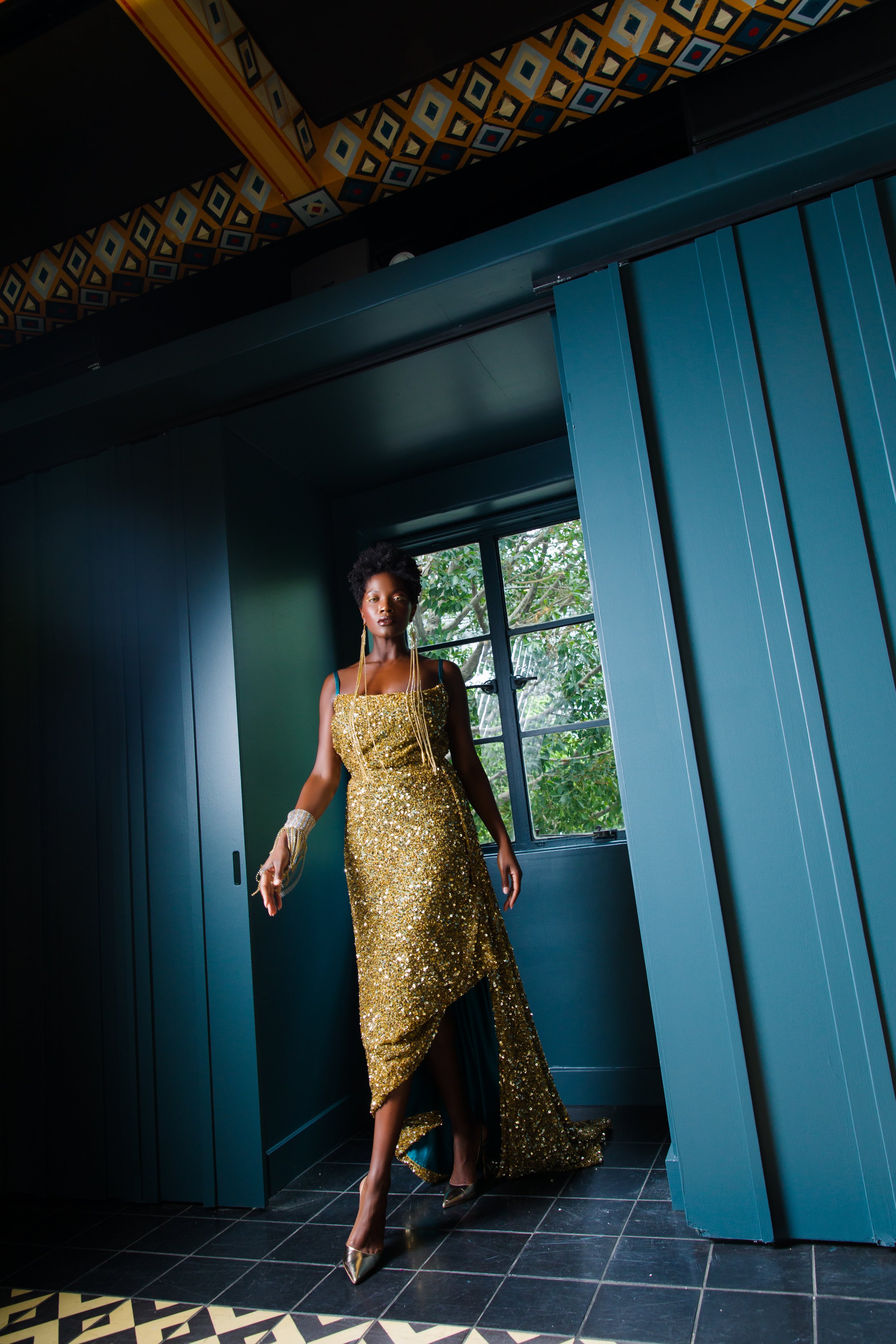  The Hand-Beaded Rich Gold Sequined Gown is lined&nbsp;with rich teal charmeuse with an asymmetrical&nbsp;draped back and a high side slit. This look is finished with an amazing rhinestone cuff with extra long crystal fringe draping down from the cuf