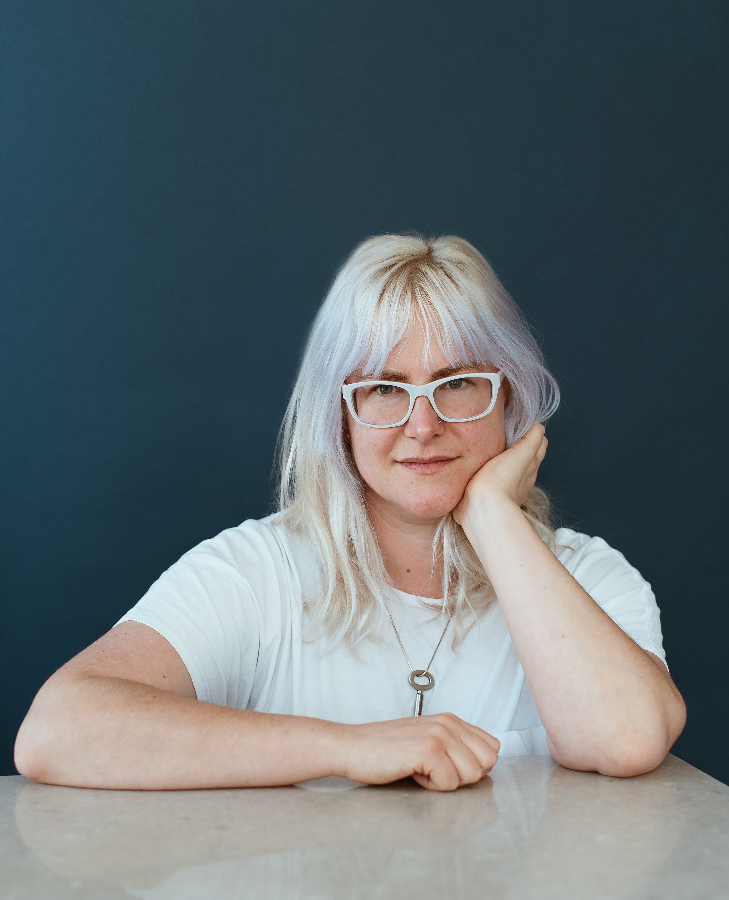  Elise, a white woman with purply-blonde hair and white glasses, looks straight at the camera. She is leaning on one hand, sitting at a marble countertop.  