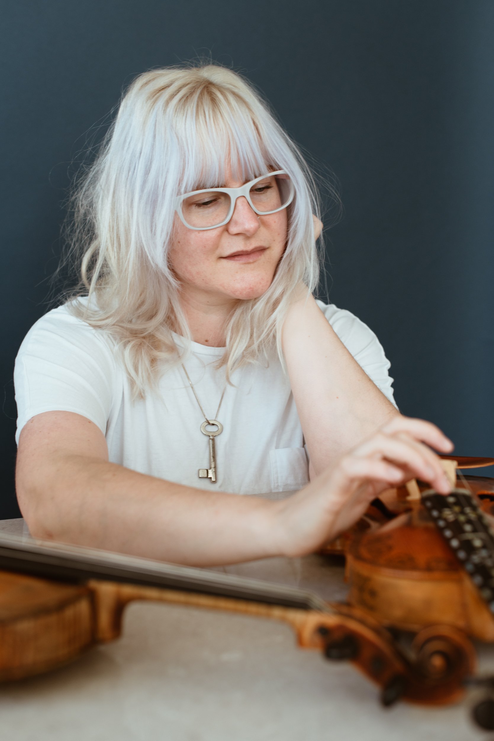  Elise, a white woman with purplish blonde hair and white glasses, touches a string on her Hardanger fiddle while looking down at it. 