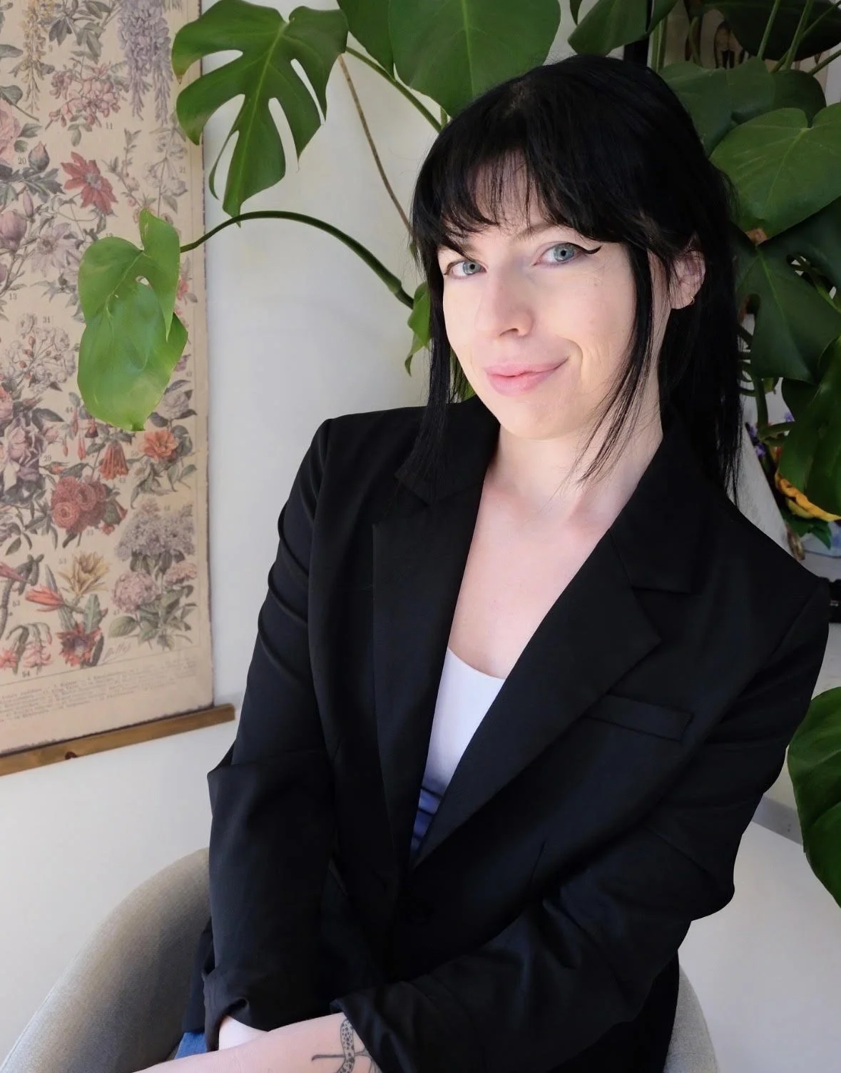A woman with long black hair, glasses, and a black shirt smiling with a colorful abstract painting in the background.