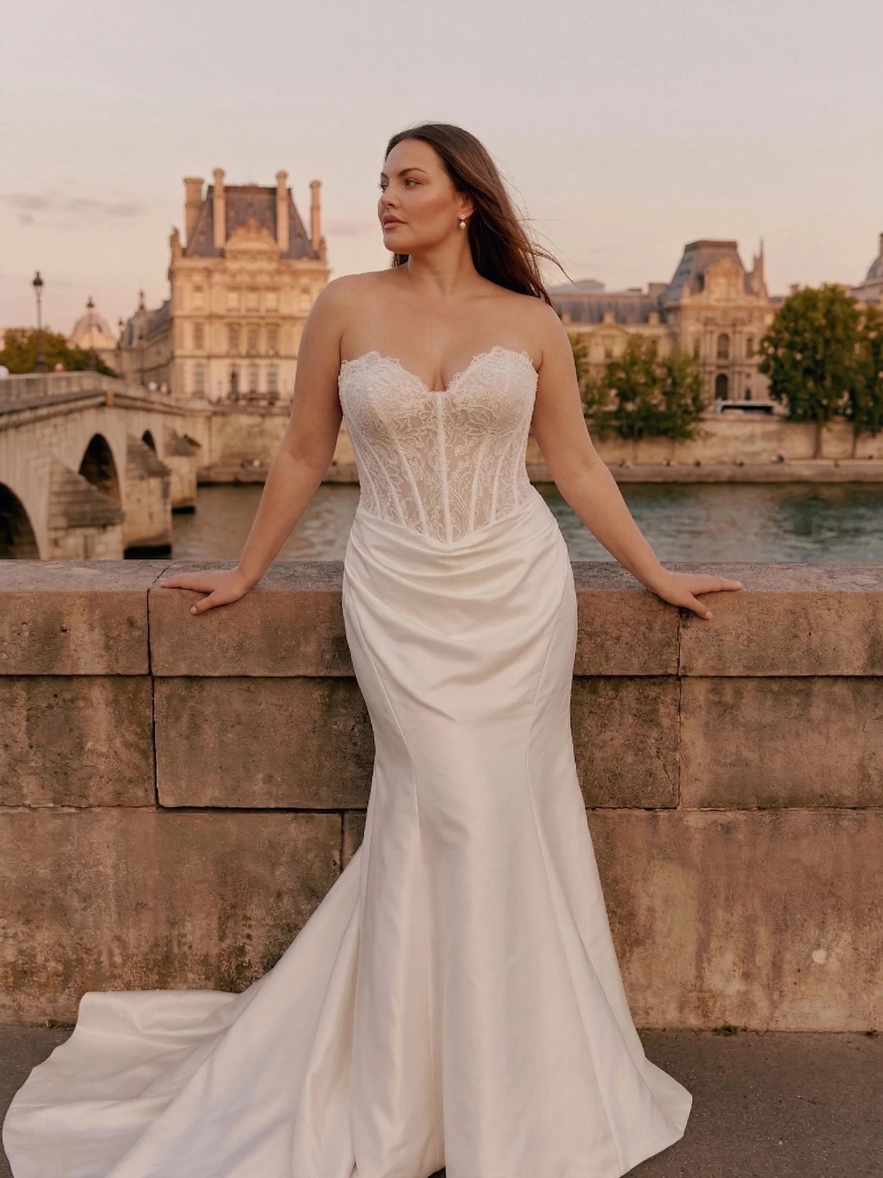 A woman in a wedding dress standing outdoors by a stone wall with historical buildings and a river in the background during sunset.