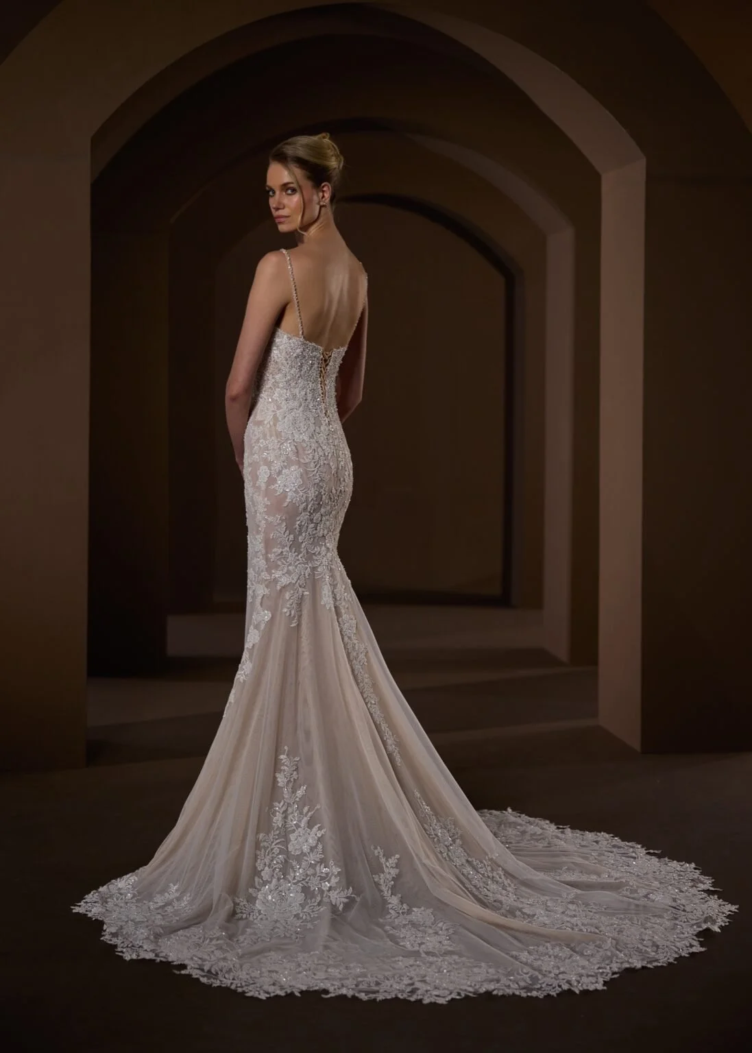 A woman wearing an elegant, fitted wedding gown with lace details, a train, and thin straps, standing in a dimly lit, arched hallway.
