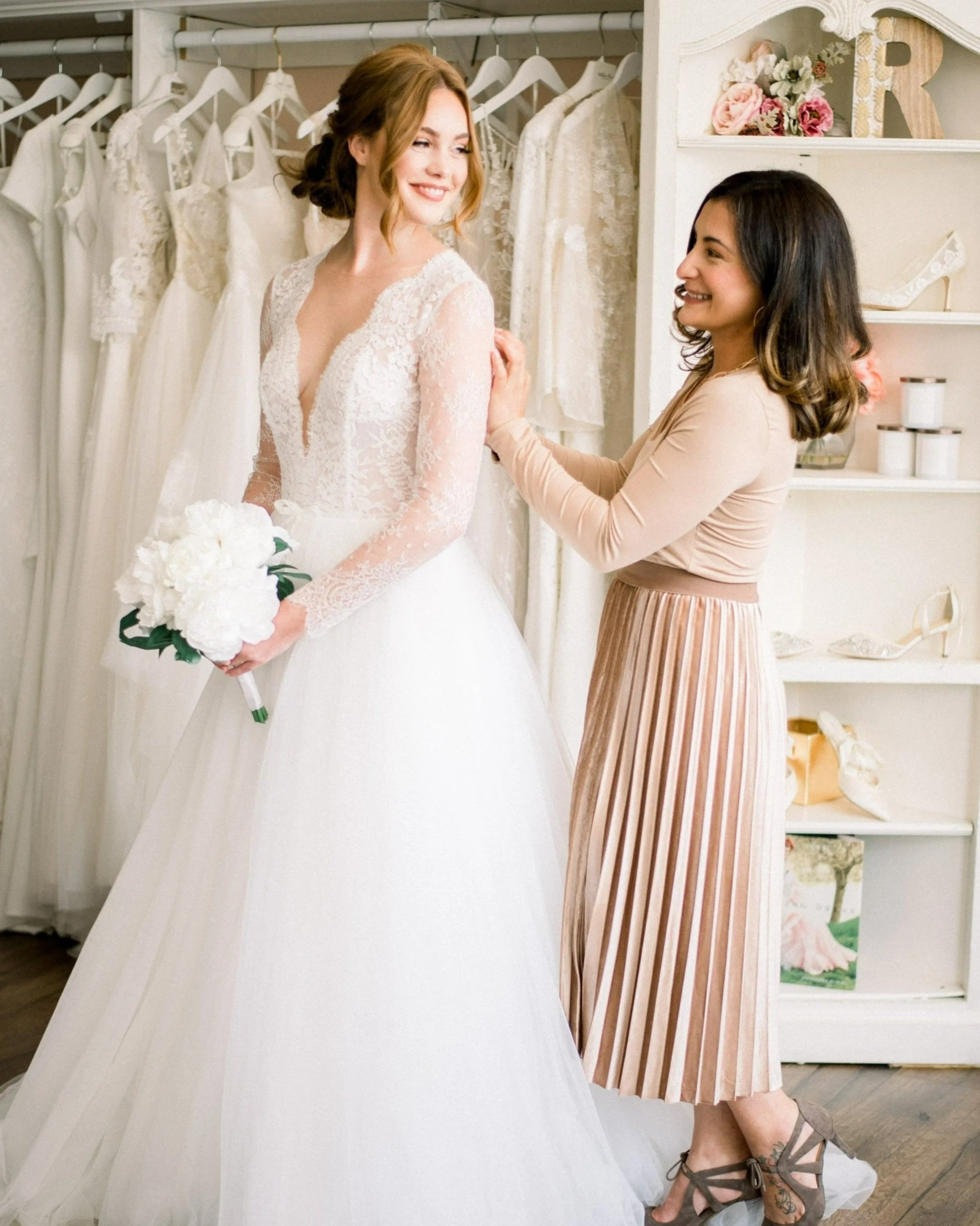 A bride wearing a lace wedding dress holding a bouquet of white flowers standing in front of a rack of wedding dresses, smiling at a woman who is adjusting her wedding dress.