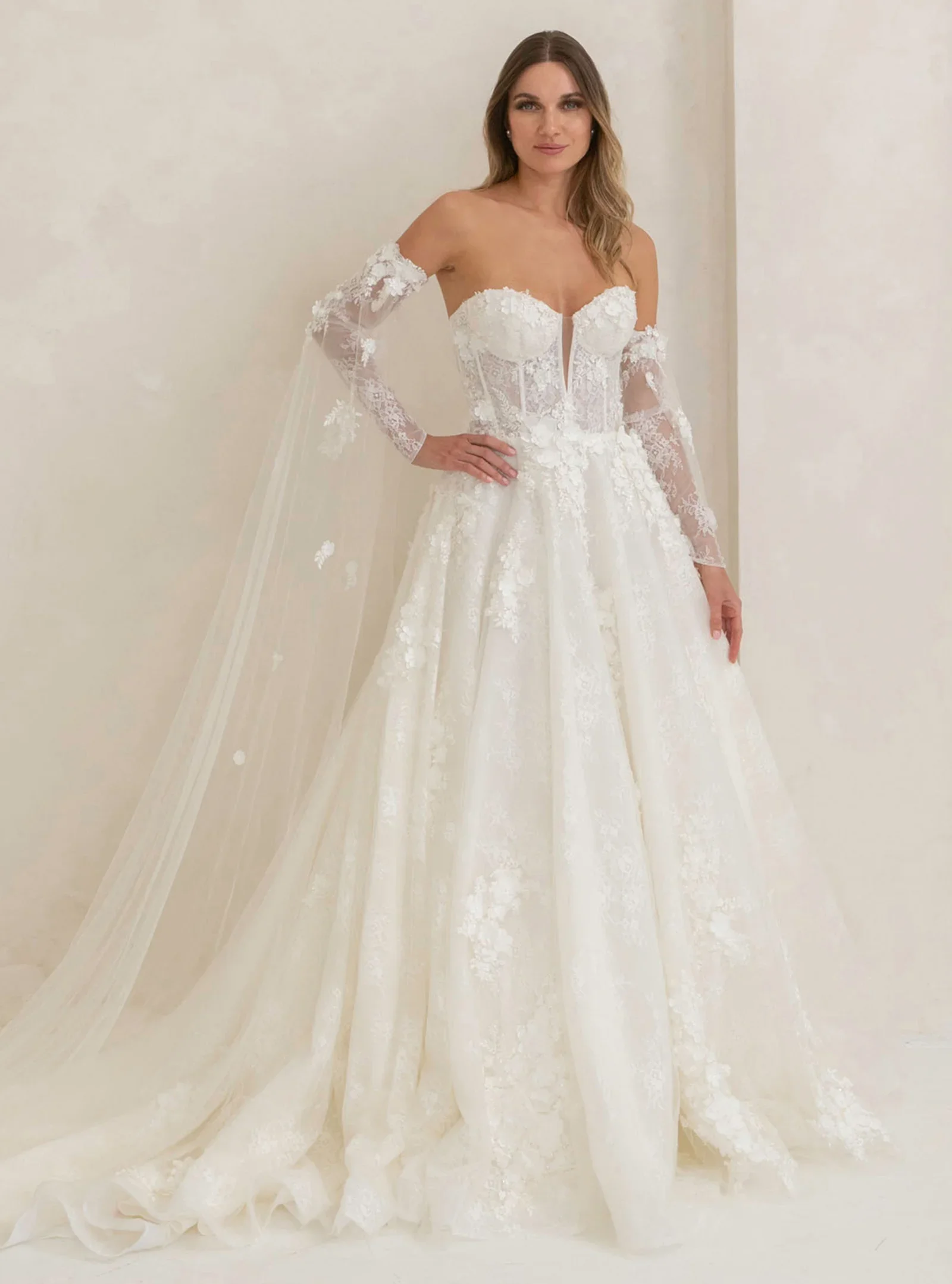 A woman in a white lace wedding gown with off-shoulder long lace sleeves, standing against a plain cream-colored wall.
