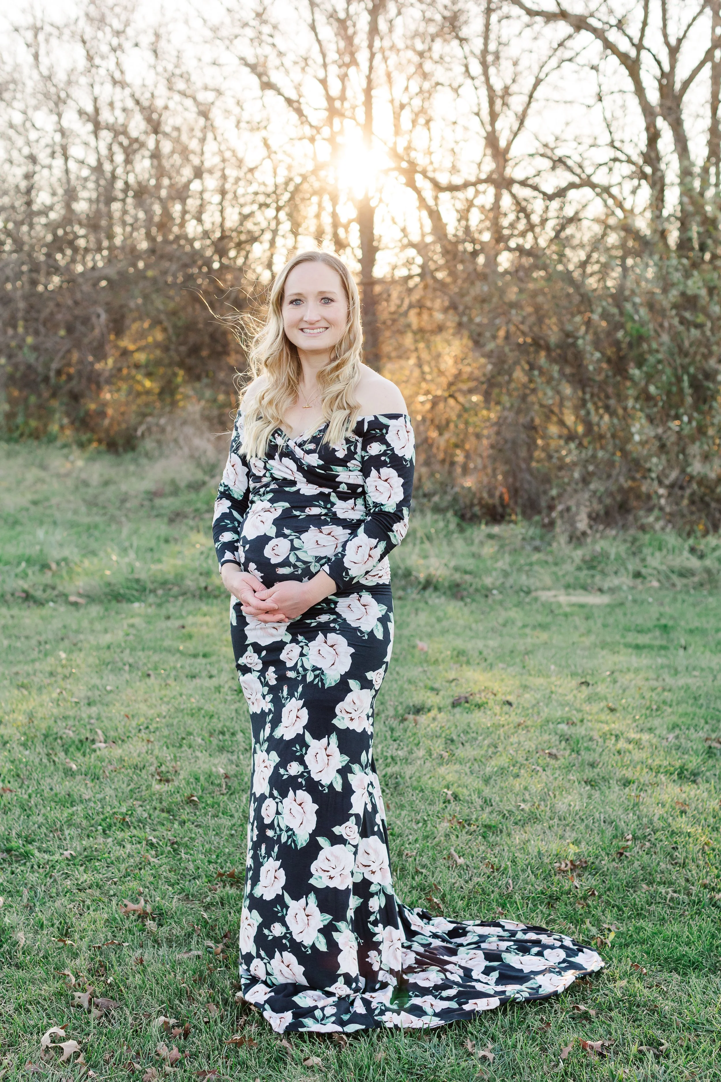 Pregnant woman in floral dress standing in a field with trees and sunset background for maternity photos