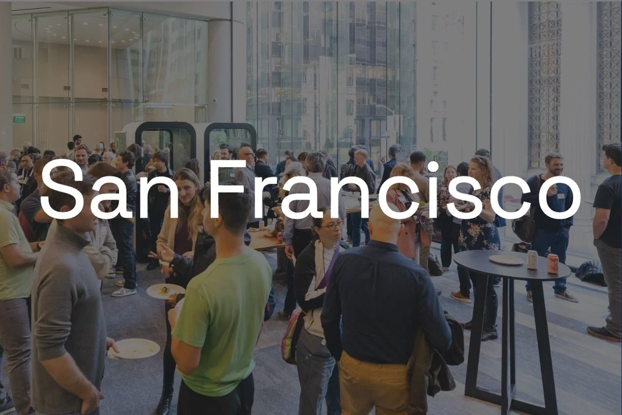 Interior of a modern lobby with a large glass window, a central seating area with benches and chairs around a tree, decorative lighting, and a view of city buildings outside. Text overlay reads 'San Francisco'.