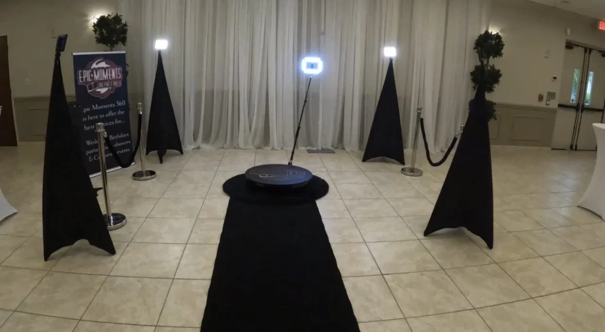 Photo of a photo booth setup with a black carpet leading to a black circular platform equipped with a ring light and a camera. The background features white curtains, and there are trees with lights on top of black stands on either side, as well as a signage banner that reads "Epic Moments 360."