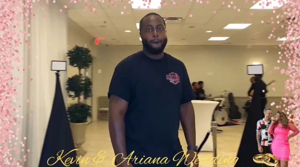 A man dressed in casual black t-shirt standing in a decorated wedding venue with pink floral borders and text that reads 'Kevin & Ariana Wedding' and a small image of a couple in wedding attire.