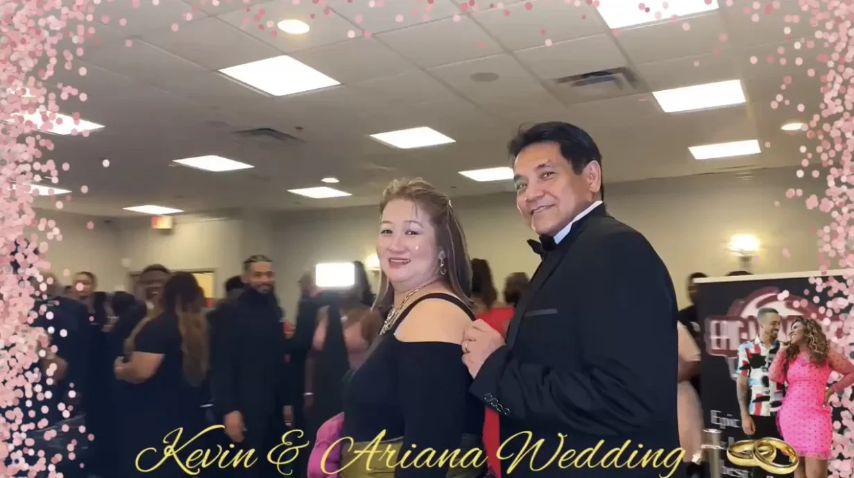 A couple at a wedding reception, the man in a black tuxedo and the woman in a black dress with cut-out shoulders smiling, surrounded by other attendees in formal attire. The photo is framed with pink flower accents and has the text "Kevin & Ariana Wedding" with wedding rings at the bottom.
