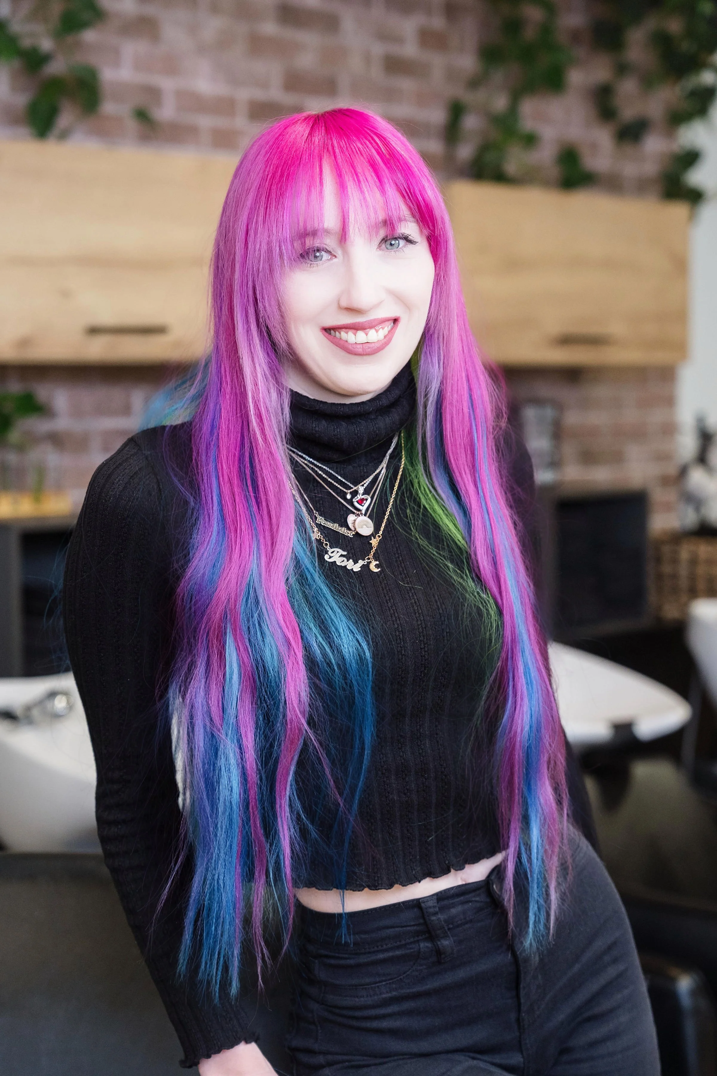 A woman with long, rainbow-colored hair and a tattoo on her arm, smiling and sitting on a couch.
