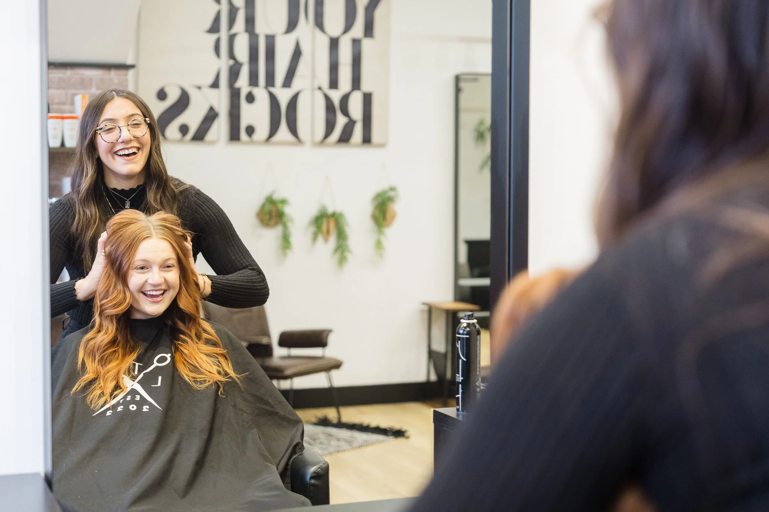 Two women, a stylist and a client, in a hair salon, with the stylist cutting and styling the client's red hair. The client, seated in a salon chair, is smiling and wearing a black cape. The stylist, standing behind her, is smiling and wearing glasses and a black sweater. The salon has a modern decor with plants on the wall and artwork in the background.