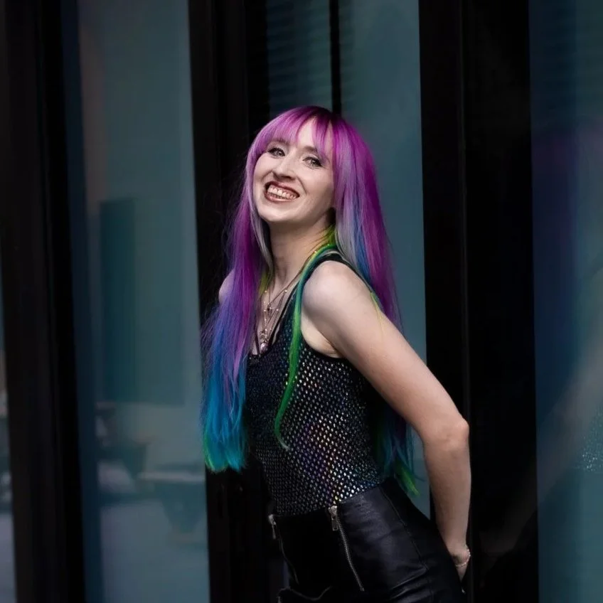 A woman with long, rainbow-colored hair and a tattoo on her arm, smiling and sitting on a couch.