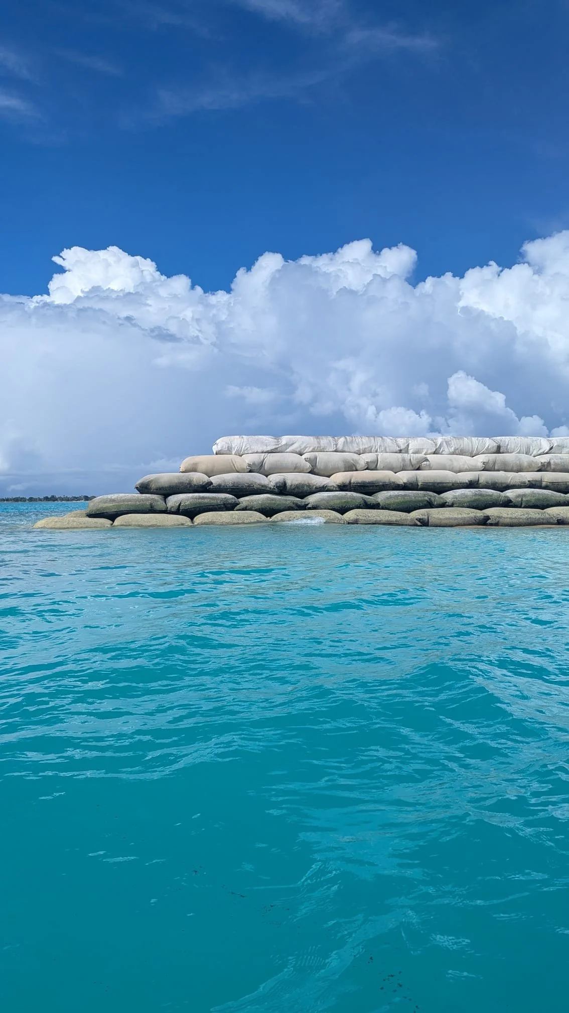 A seawall made of large geotextile bags on a body of water under a partly cloudy sky.