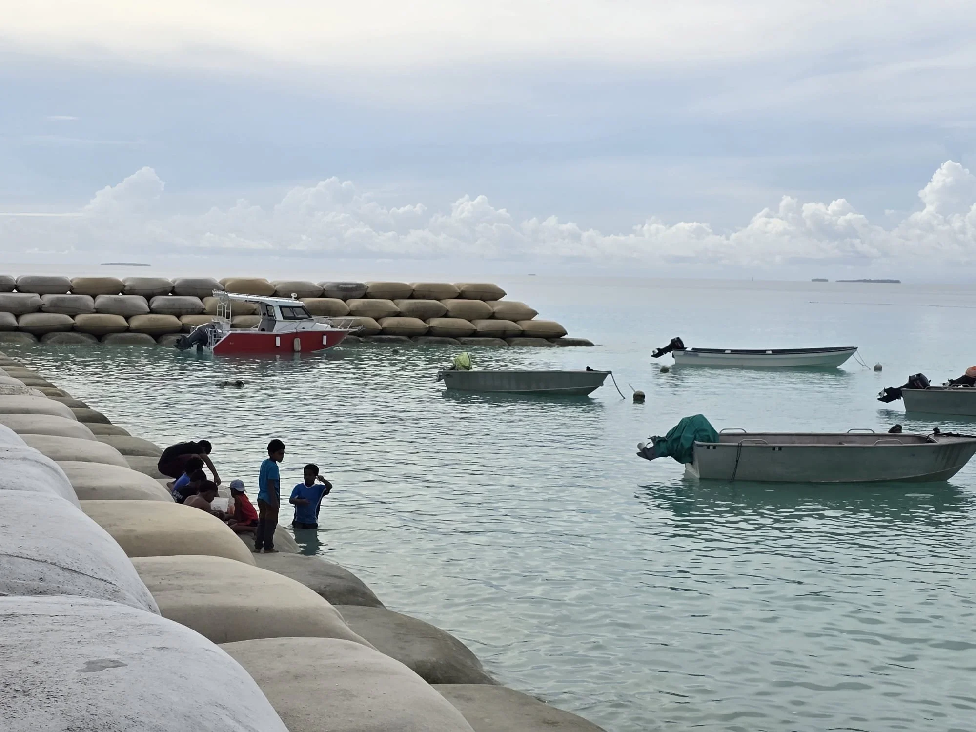 Children playing and sitting along a breakwater by calm sea, with boats moored nearby and cloudy sky overhead.