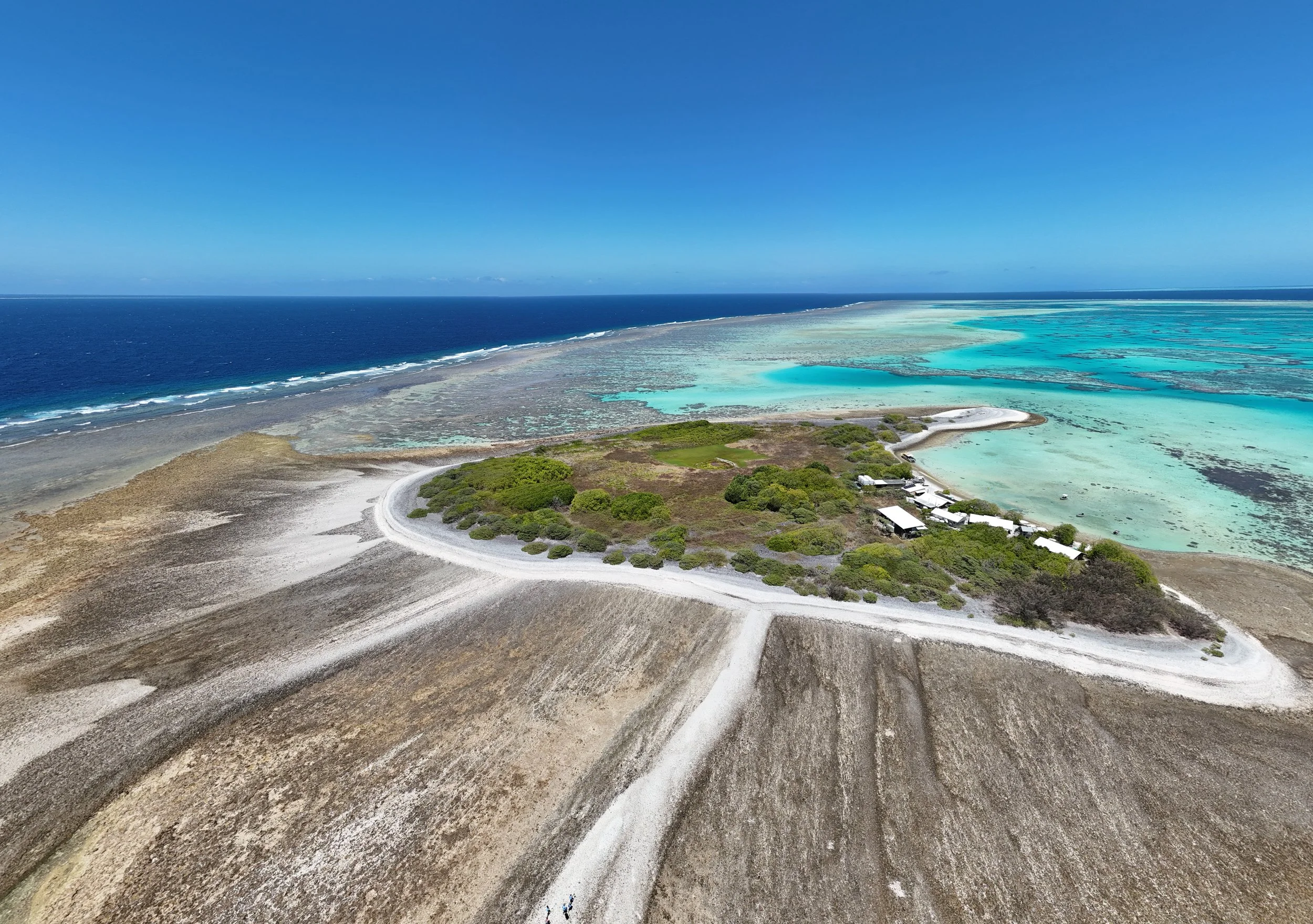 Aerial view of a coastal landscape with a sandy shoreline, turquoise and blue water, some green vegetation, and a clear blue sky.