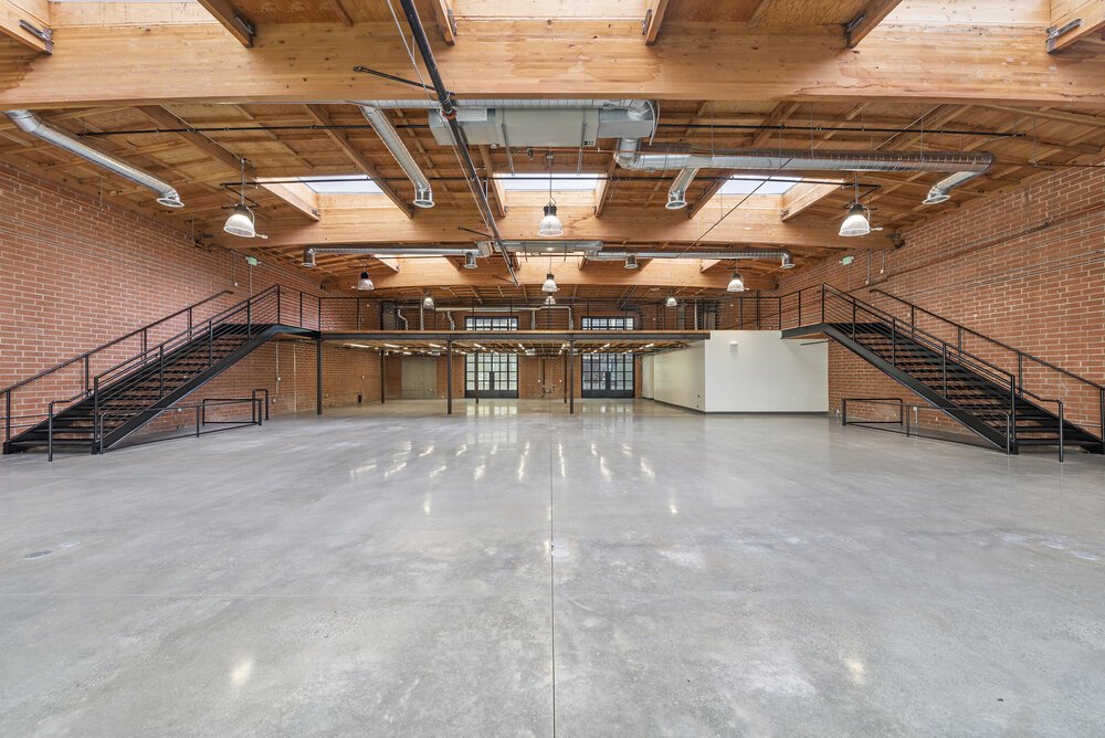 Empty industrial-style loft with red brick walls, wooden ceiling with exposed ductwork, and concrete floor. Two black staircases with railings leading to a mezzanine level.