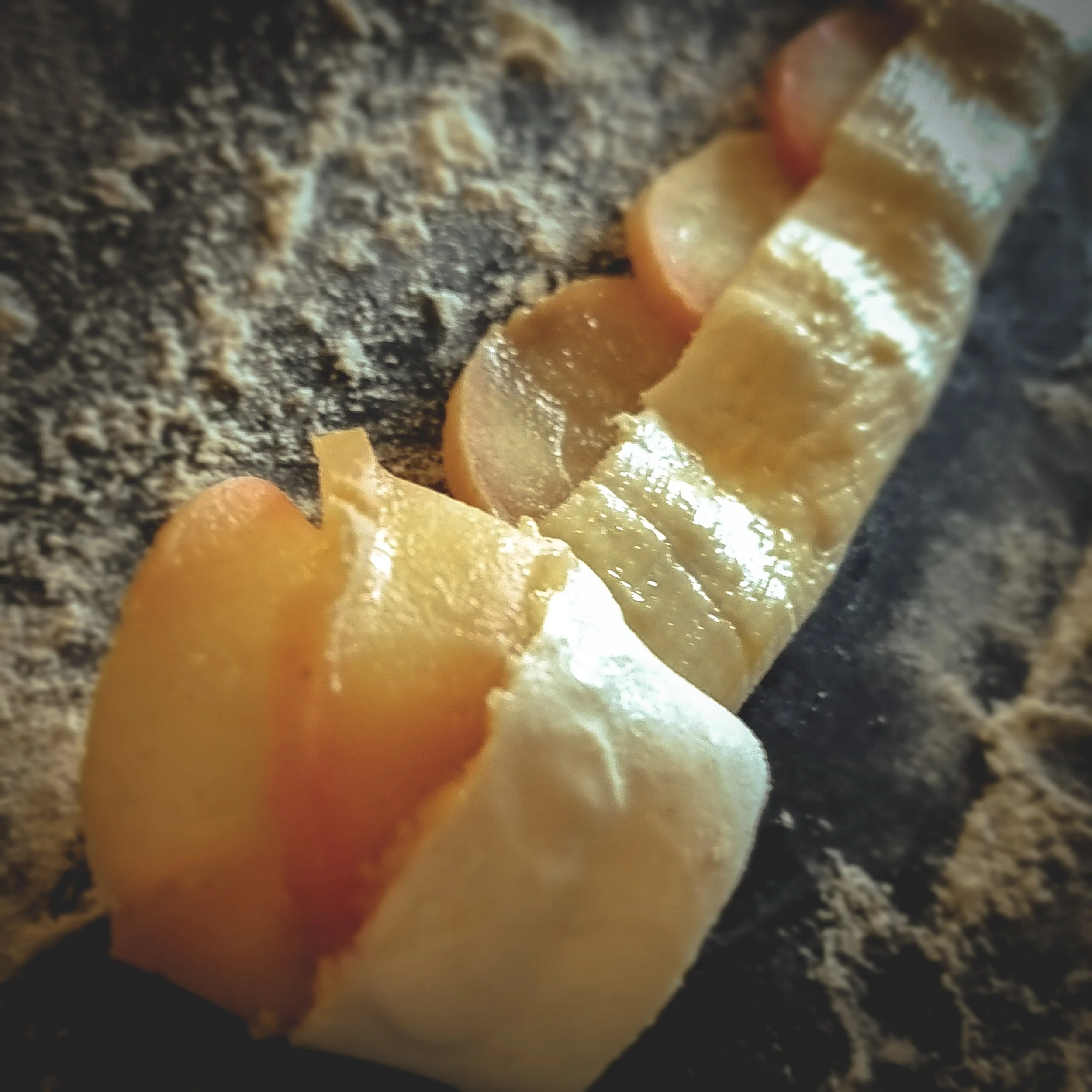 Apple slices lain along the long edge of a pastry strip which has been folded up over the apples and is being rolled gently to form a rose on a black marble pastry board dusted with flour