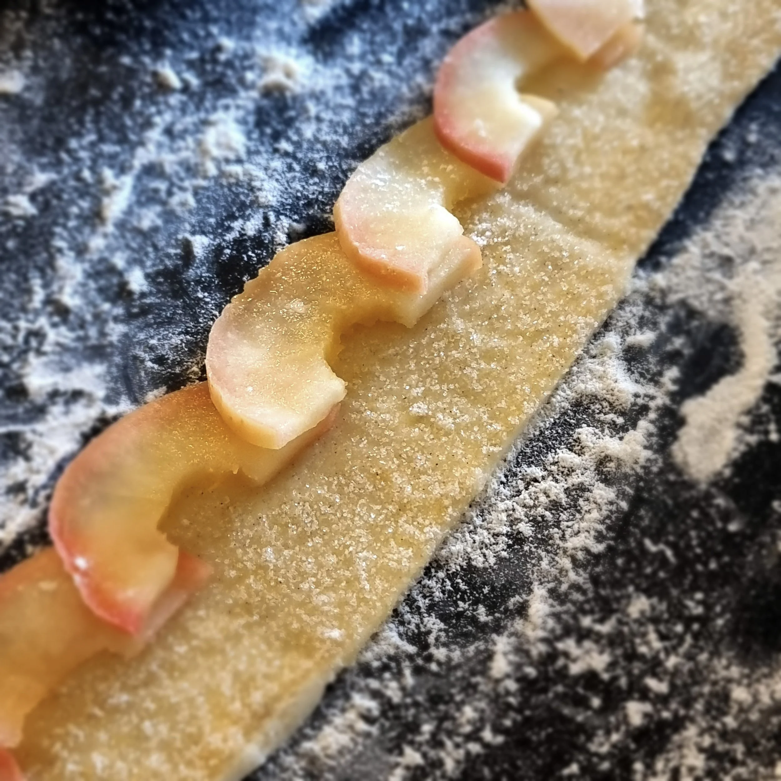 apple slices laid along the long edge of a strip of pastry which has been brushed with melted butter and sugar on a black marble pastry board dusted with flour