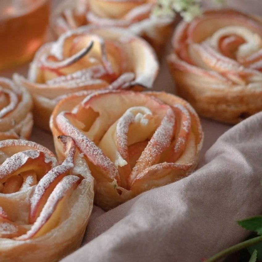 Apple Roses sit in a basket lined with linen fabric, puff pastry is c golden and the apple petals are dusted with icing sugar,