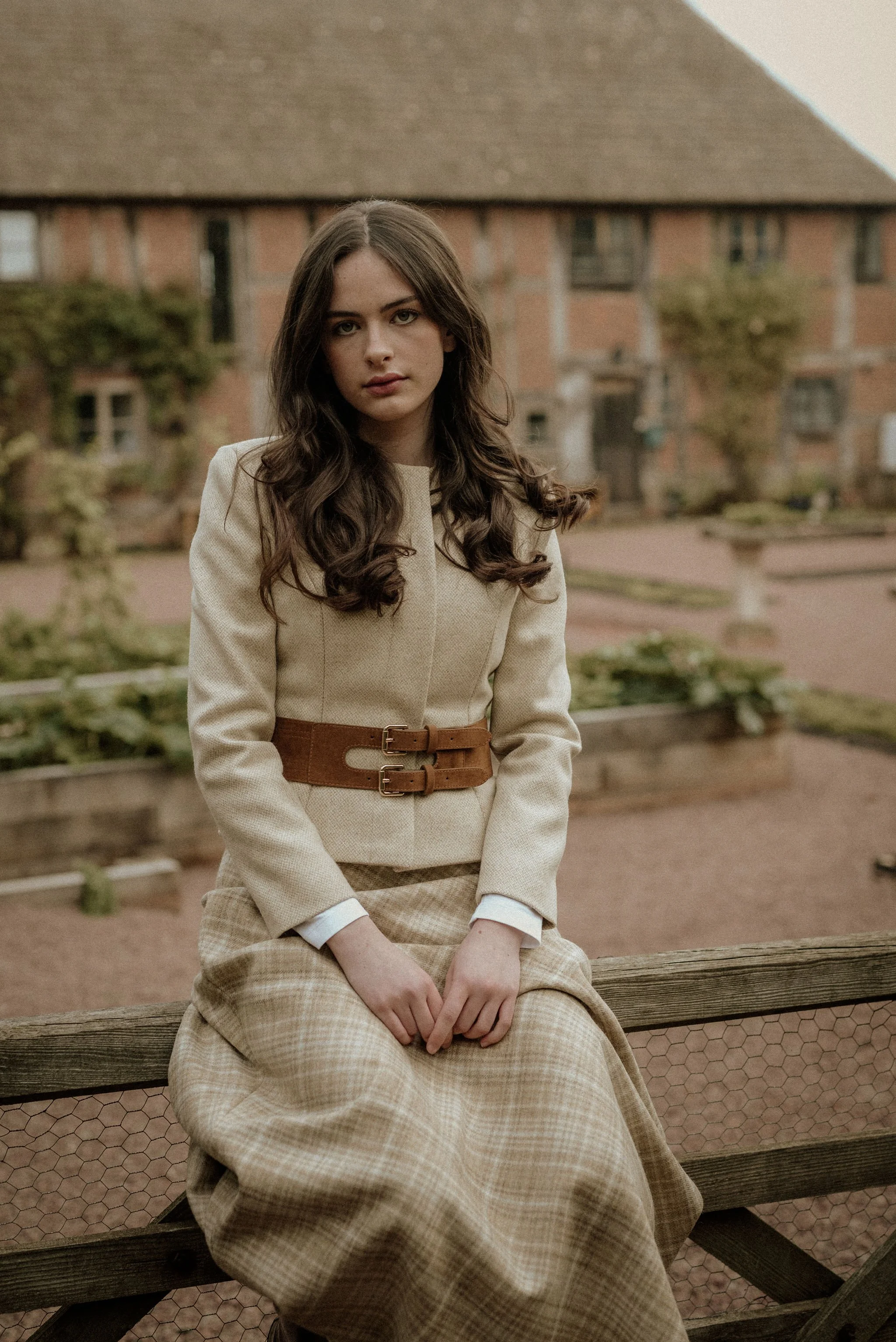A young woman with long, wavy brown hair sits on a wooden farm gate outside, wearing a tweed jacket with a wide brown double buckle belt and a plaid skirt, with a blurred brick building and vegetable garden in the background.