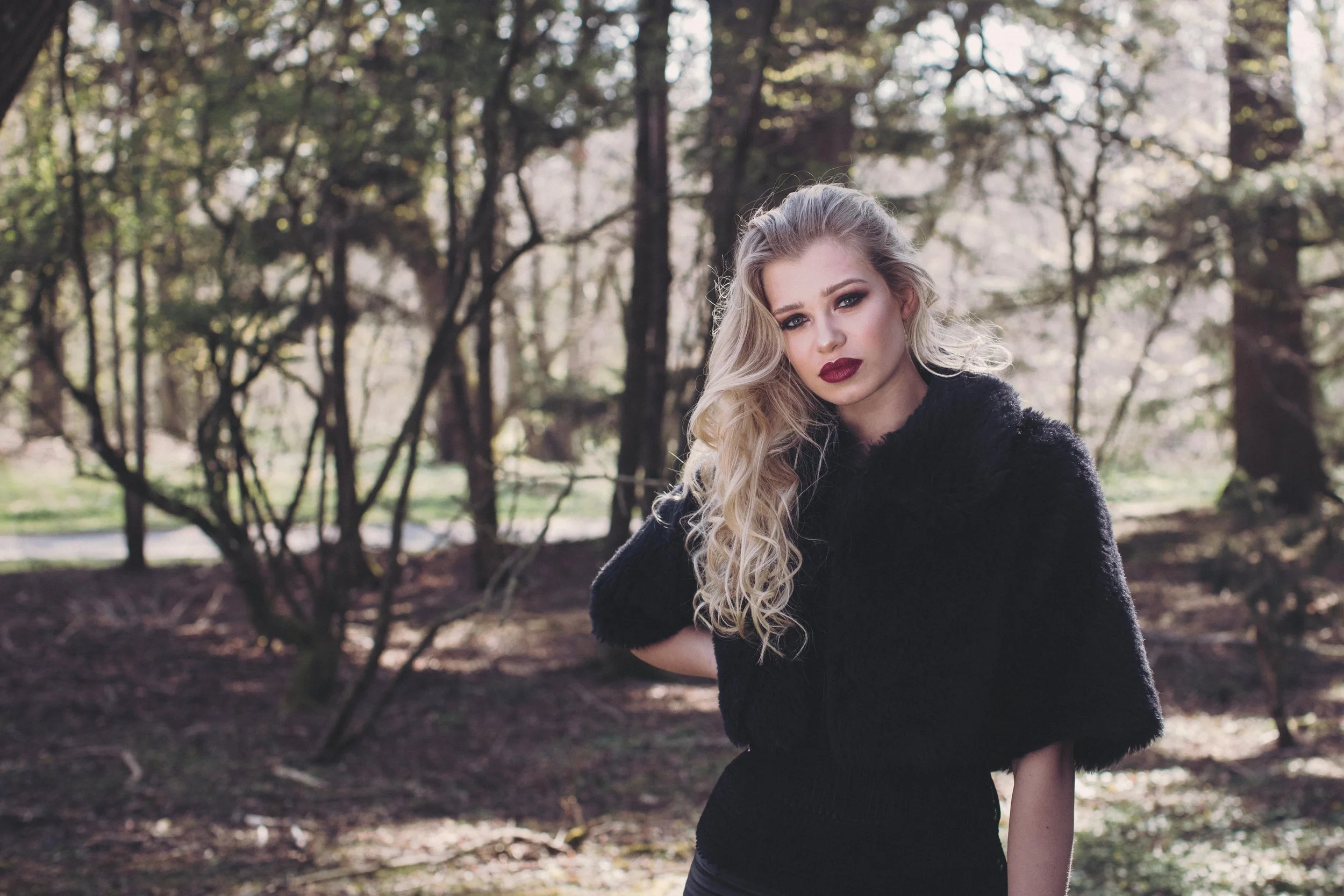 A young woman with long blonde curly hair and evening makeup, wearing a black natural fibre faux fur capelet, standing outdoors in a wooded area with trees and a pathway in the background.