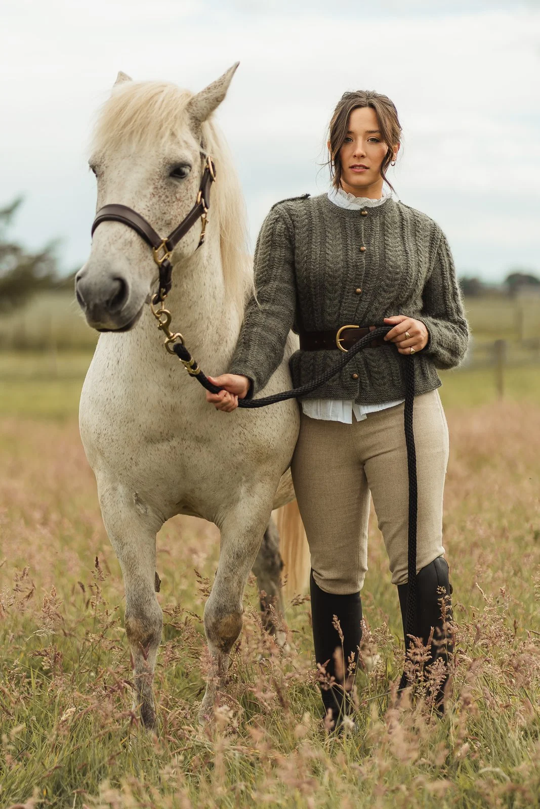 A woman standing next to a white horse in a grassy field wearing a cable knitted jacket with belt and wool trousers, holding the horse's reins, with a cloudy sky in the background.