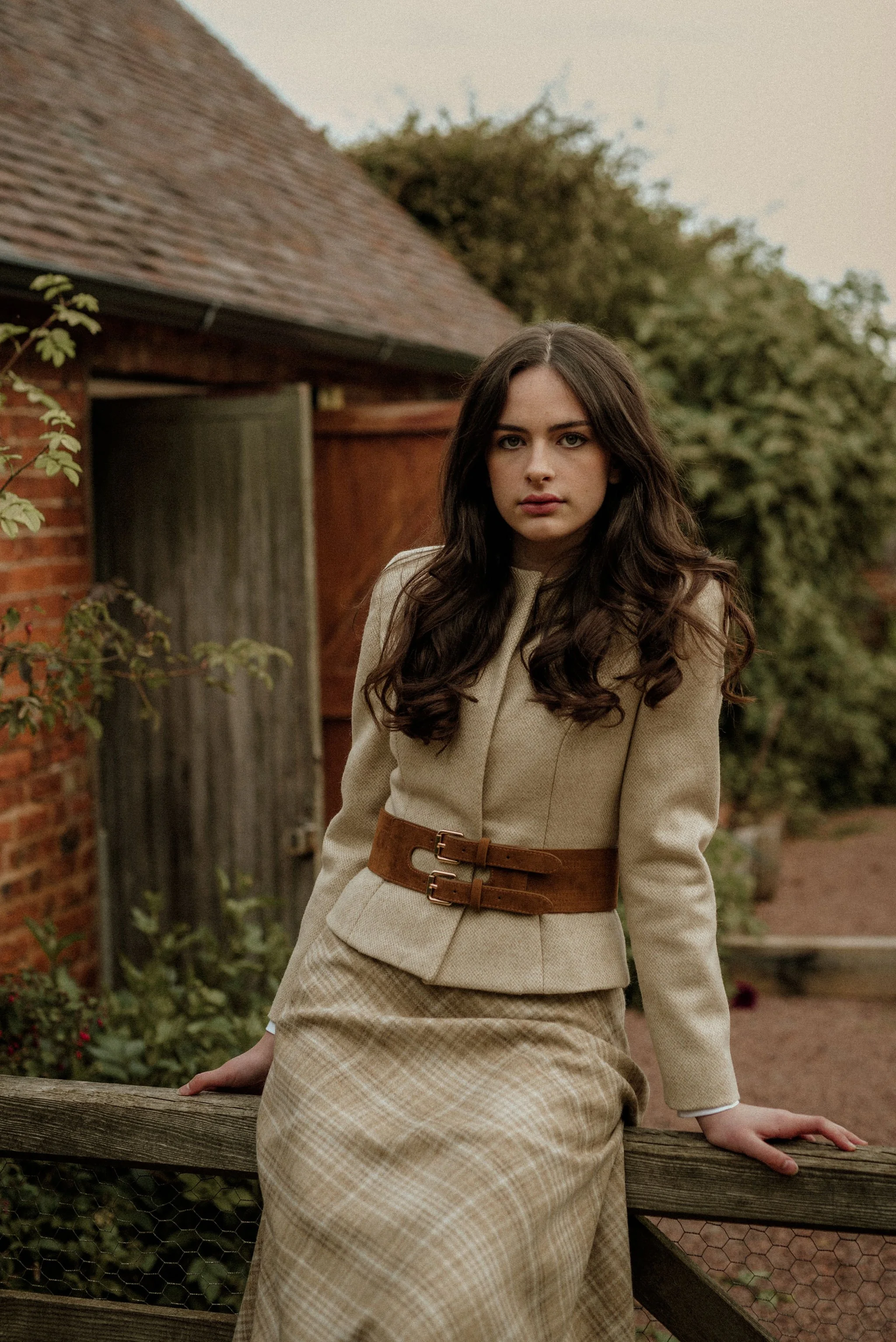 A young woman with long dark hair wearing a beige tweed jacket with a wide brown double buckle belt and a matching plaid skirt, sitting on a wooden farm gate outdoors near a brick and wooden building with greenery in the background.
