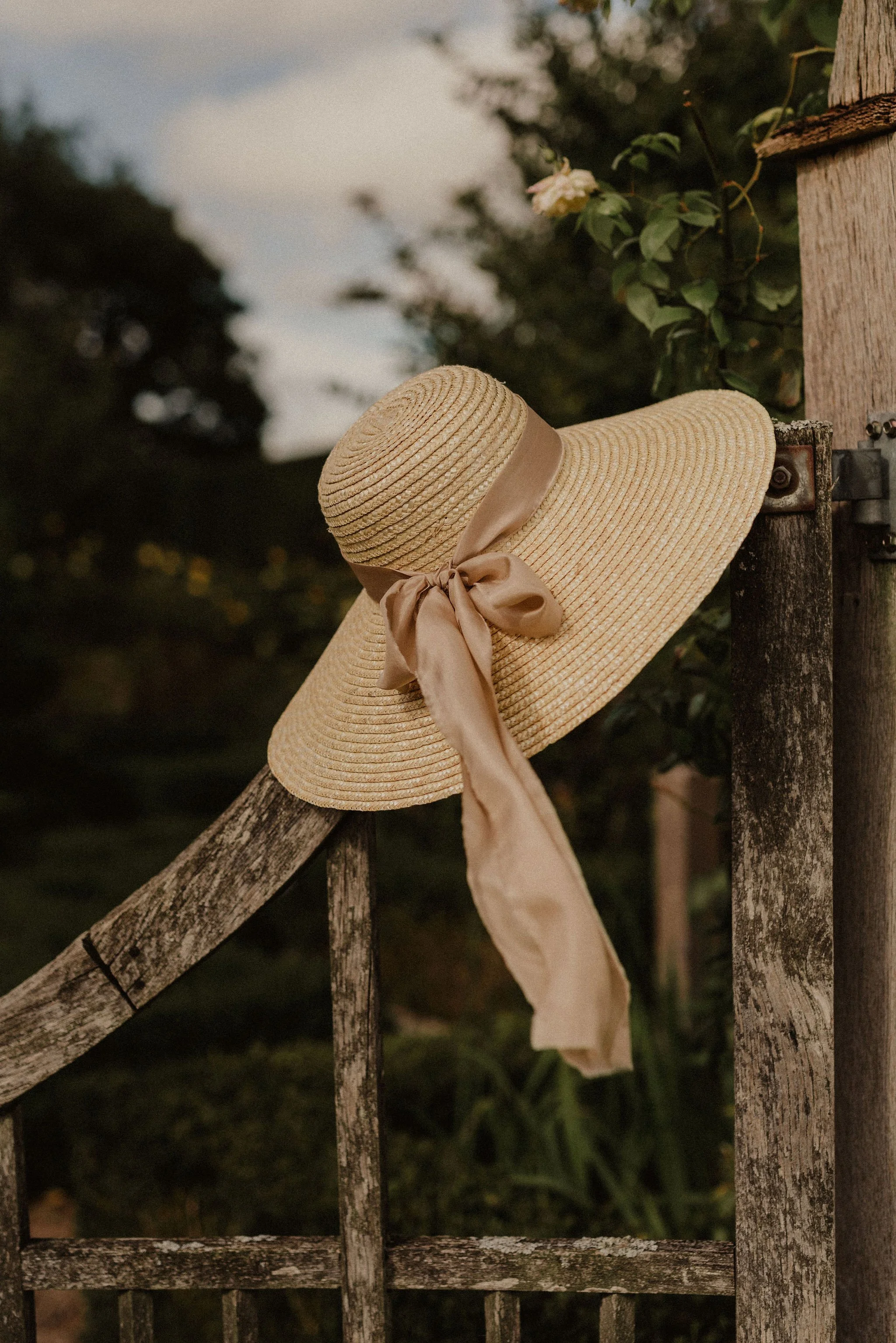 A beige straw sunhat with a satin ribbon tied in a bow, hanging on a weathered wooden fence.
