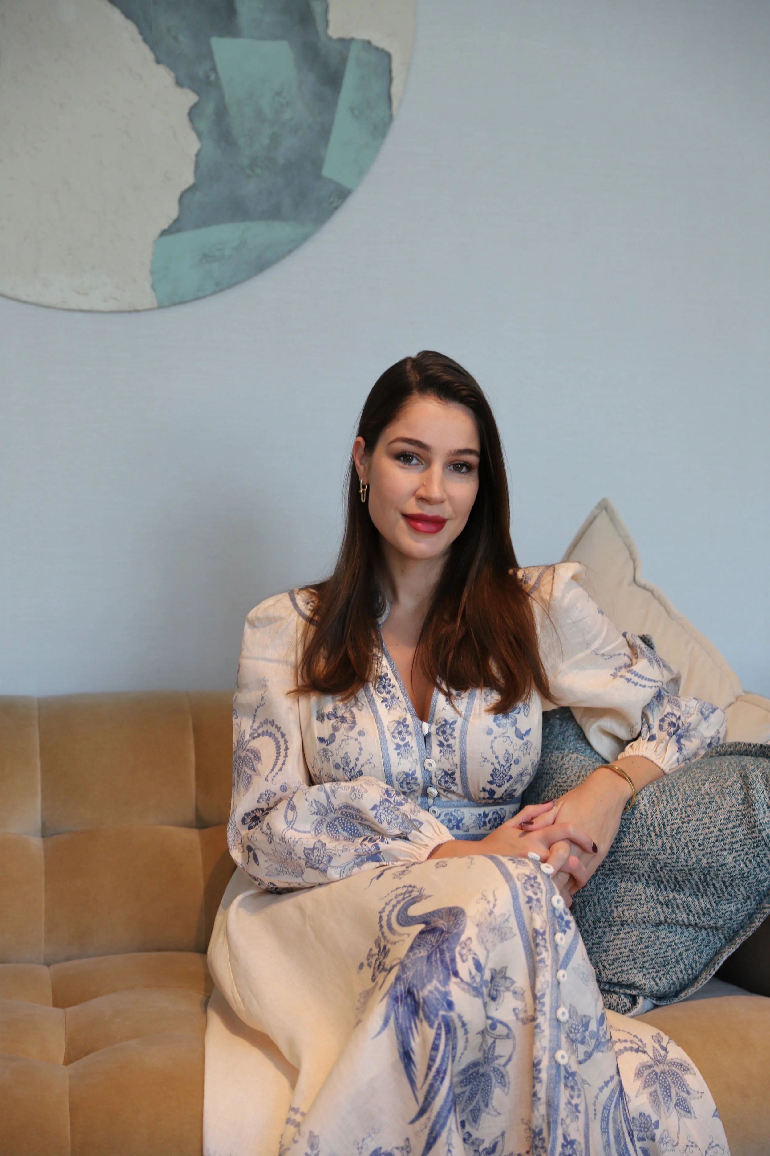 A young woman with long brown hair, wearing a white and blue patterned dress, sitting on a beige sofa with a cushion, in a room with a light blue wall, and a framed world map hanging above her.