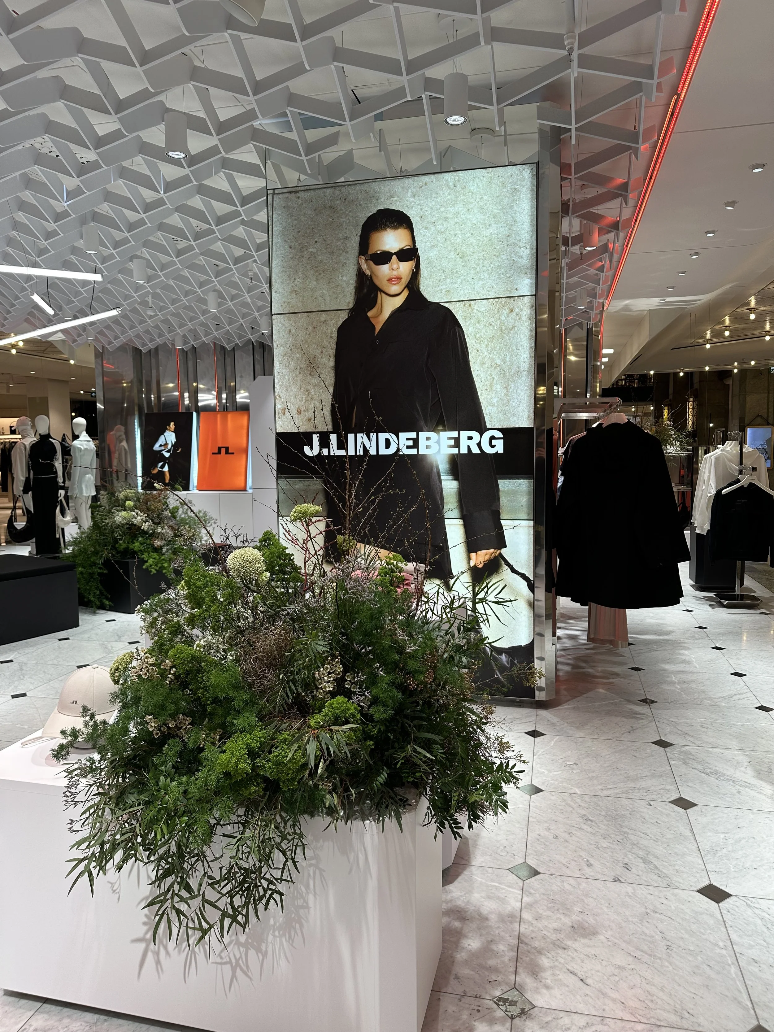 A fashion store display with a large advertisement featuring a woman wearing black sunglasses and a black jacket. The store has a modern design with geometric ceiling panels, clothing mannequins, racks of clothes, and a floral arrangement in the foreground.