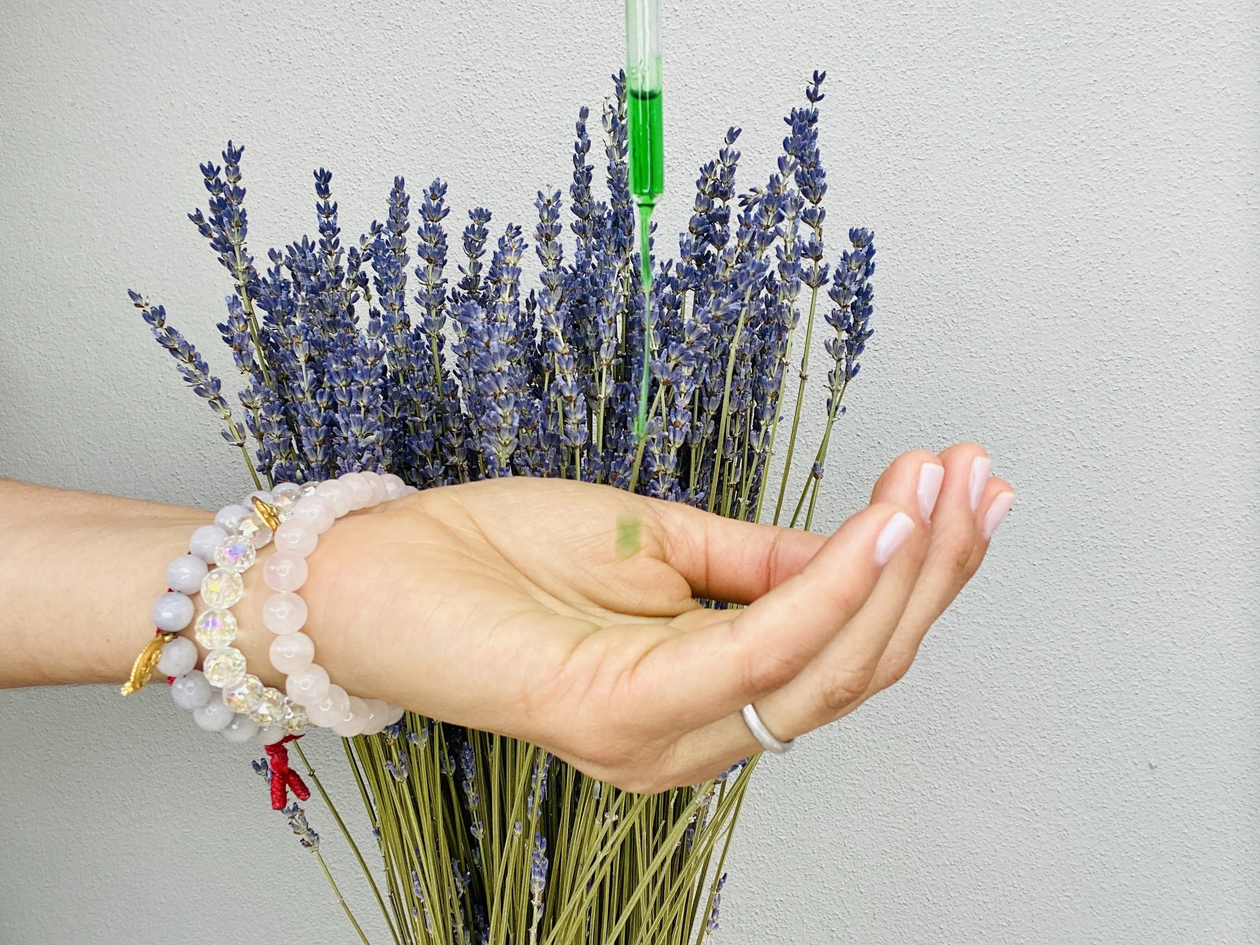 A hand with beaded bracelets receiving a green serum. Lavender flowers in the background