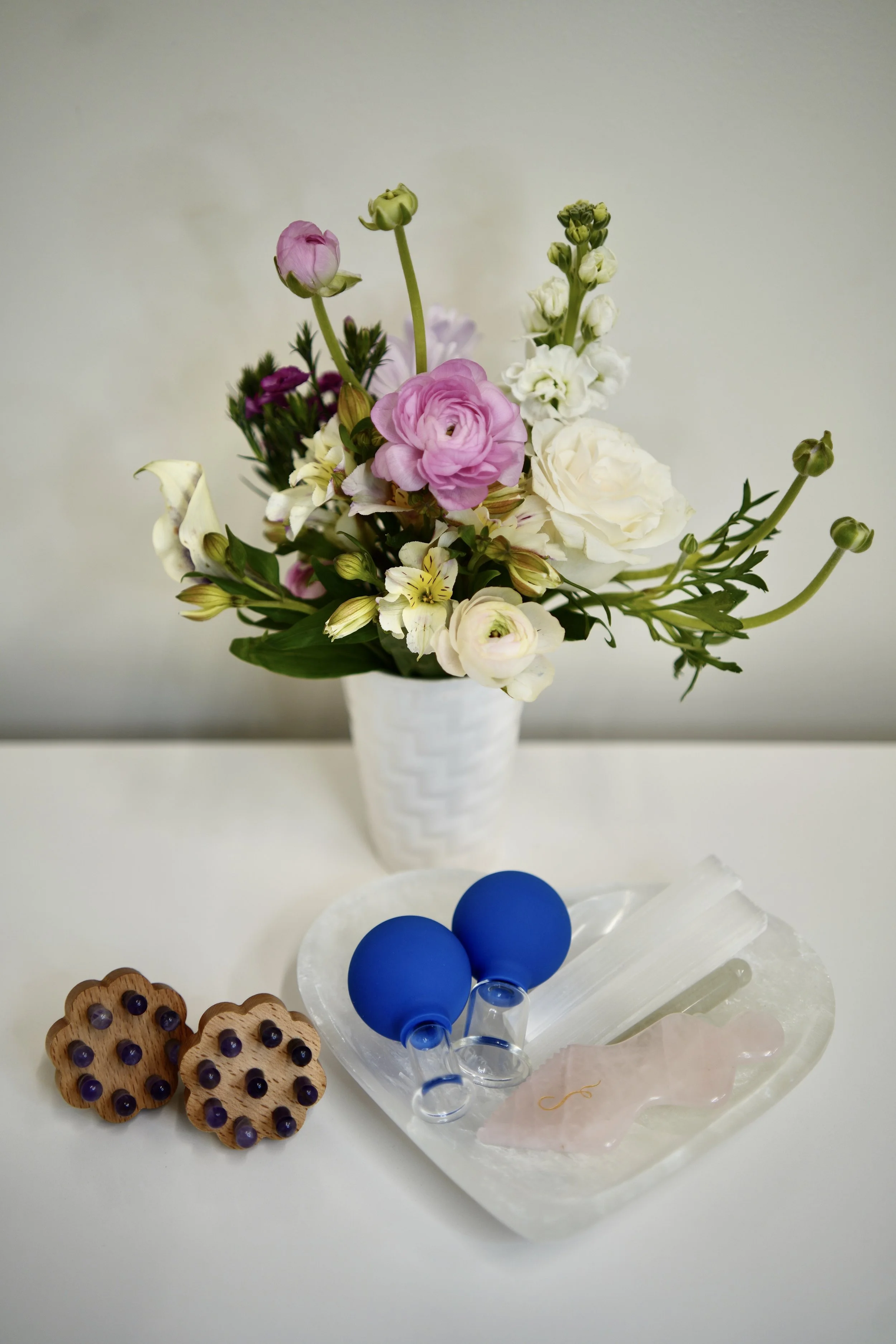 A still life of a white vase with pink and white flowers, two wooden cookies with purple dots, two blue balloons, and a pink and clear glass figurine on a white surface.
