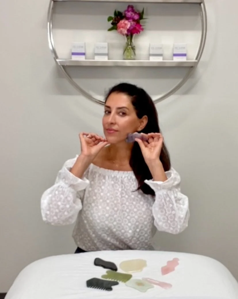 Woman demonstrating use of a gua sha tool on her face in a spa setting, with various gua sha stones displayed on a table and skincare products on a shelf in the background.
