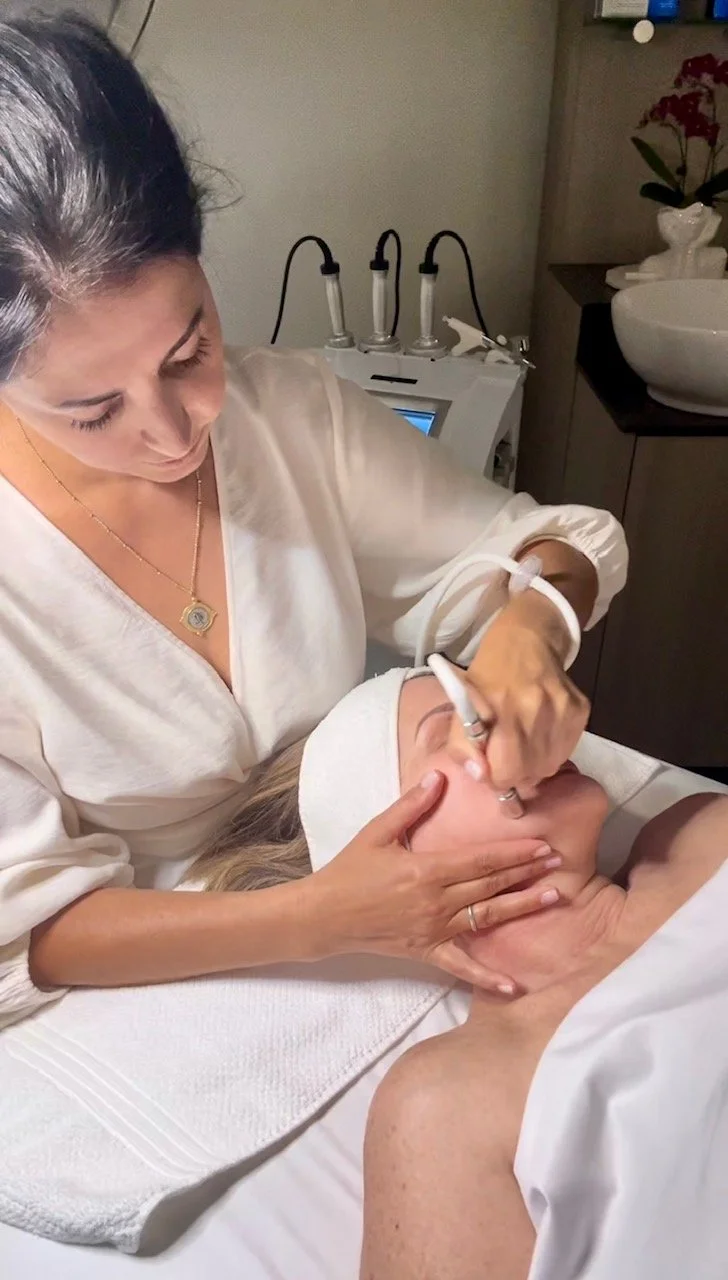 A esthetician performs a facial treatment on a woman lying down in a spa or clinic setting.