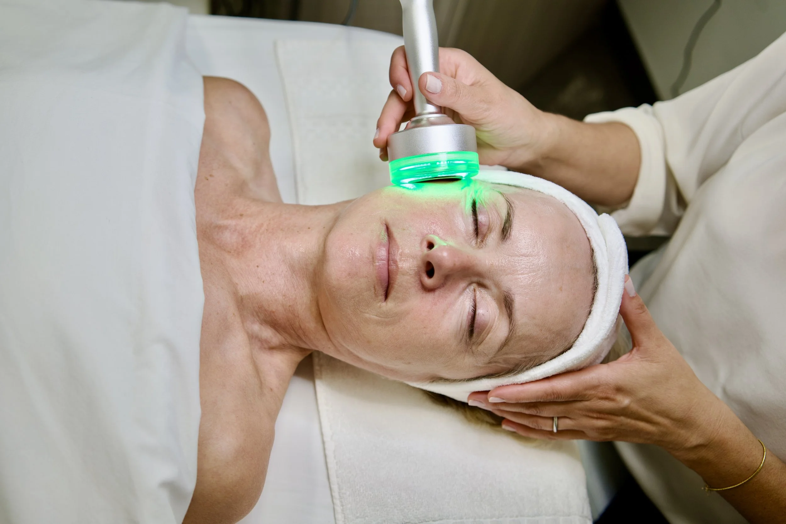 A woman receiving a facial treatment with a device emitting green light, lying on a treatment bed with her eyes closed and a towel wrapped around her head.