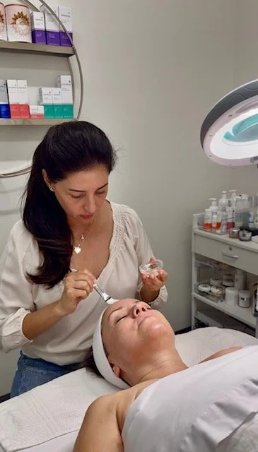 A woman receiving a chemical peel treatment from an esthetician in a treatment room with clorful boxes in the back