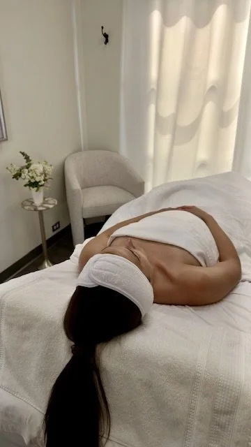 A woman lying on her side on a bed, with her head covered by a towel, in a bright room with white curtains, a white armchair, and a small table with a flower vase.