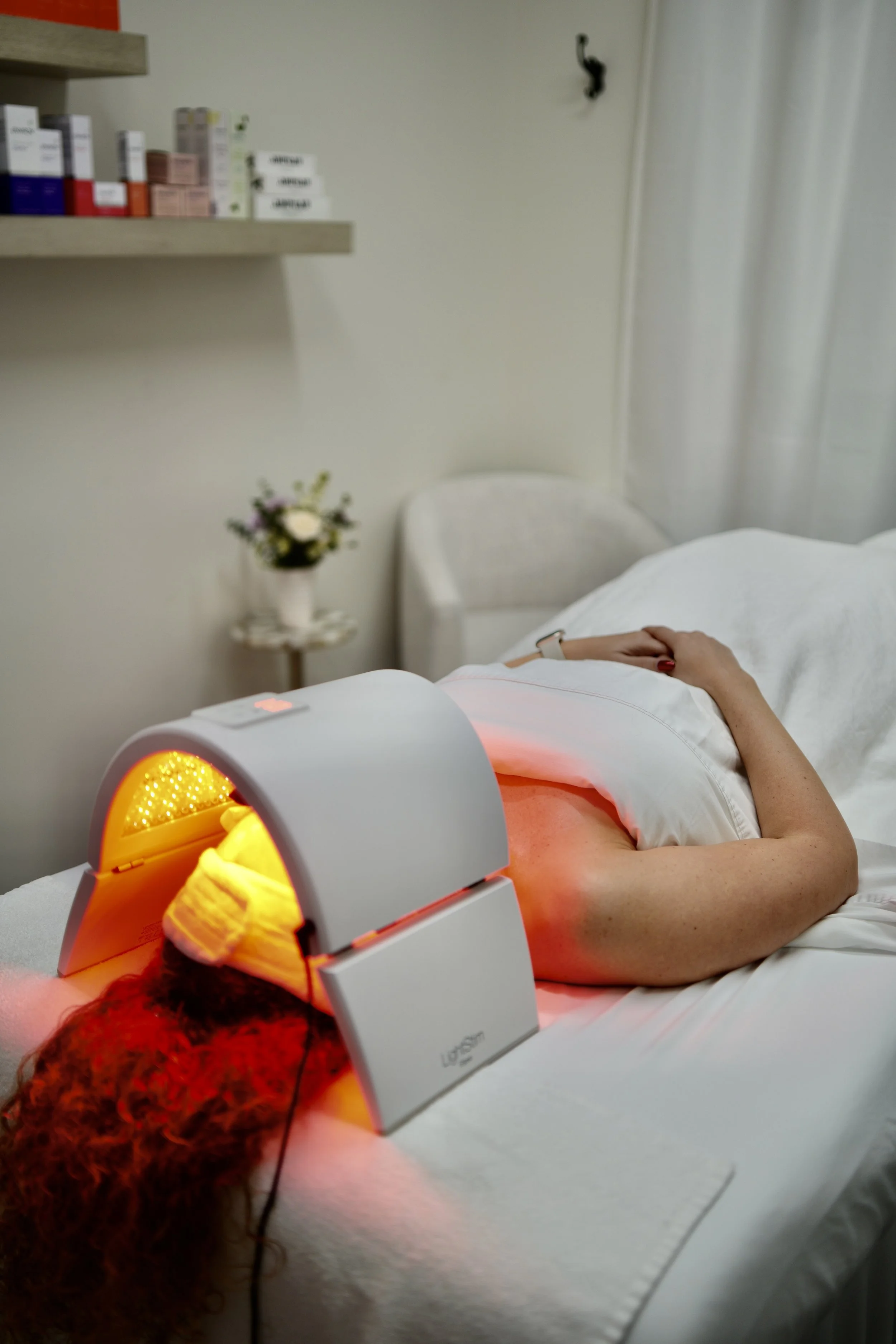 Person receiving red light therapy in a clinical setting, lying on a bed with a device emitting red light positioned over their face and chest.