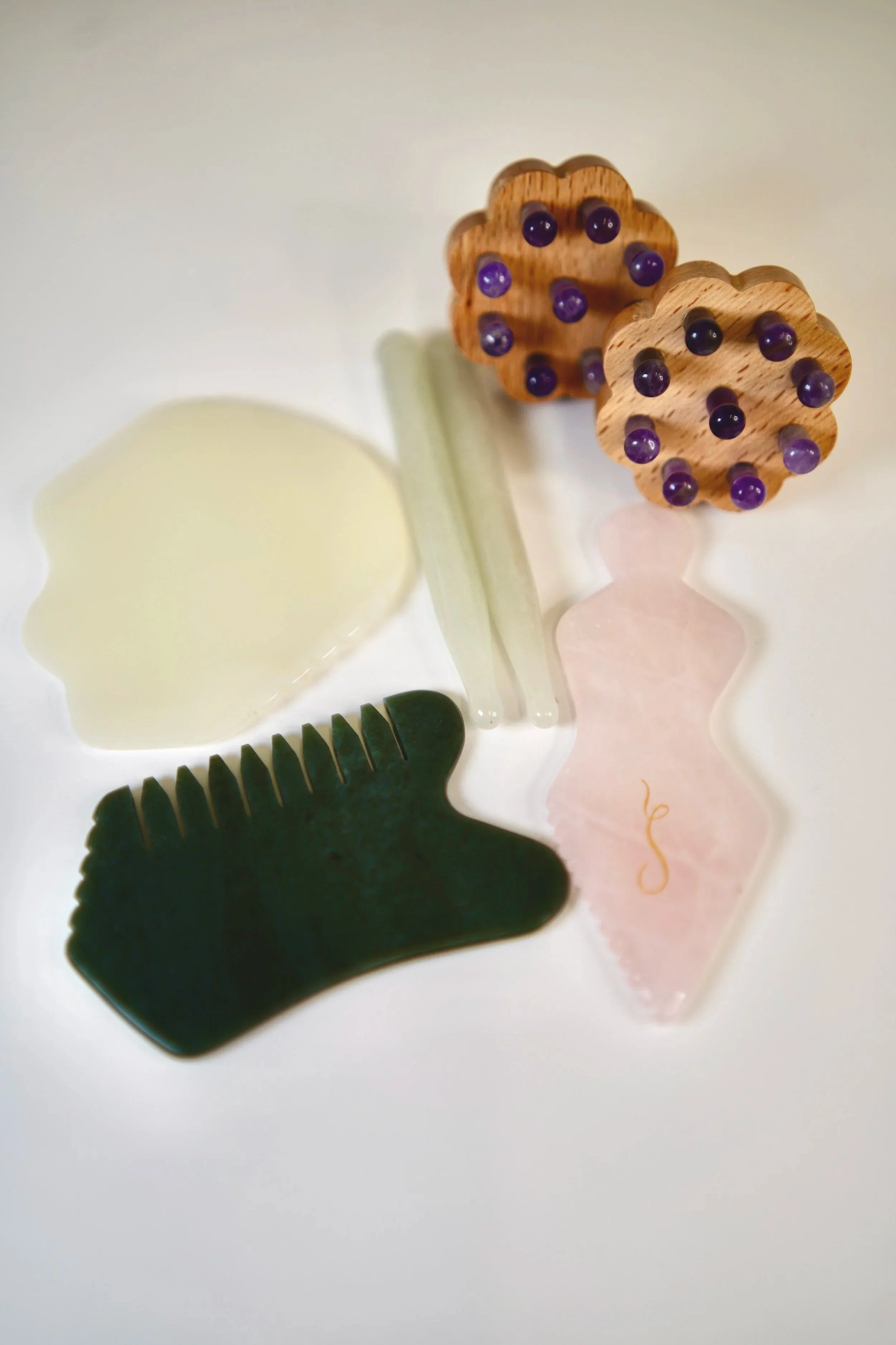A collection of different gua sha tools and wooden massage rollers with purple beads, arranged on a white surface.