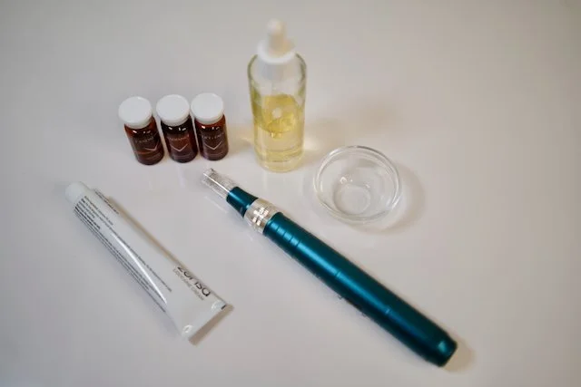 A collection of skincare or cosmetic products and tools on a white surface, including small bottles, a glass dropper bottle, a tube, a small dish, and a blue electronic device, possibly a skincare device or tool.