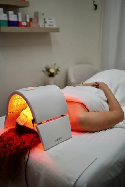 Person lying on a bed receiving red light therapy from a medical device in a clinical setting with shelves of medicine and flowers in the background.