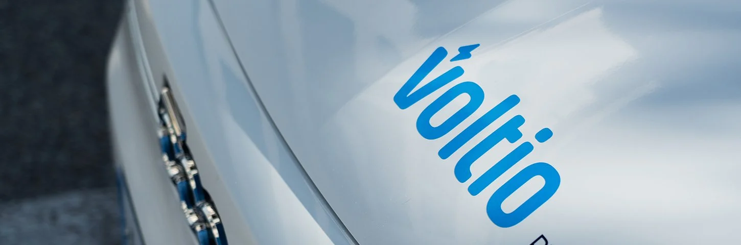 Close-up of the front part of a WooHoo vehicle with a silver or light gray exterior and blue logo, parked on a street.