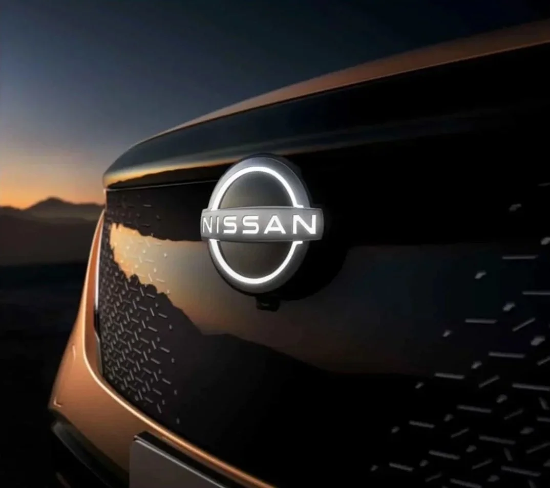 Close-up of a Nissan car emblem on the front grille, with a sunset or sunrise in the background.