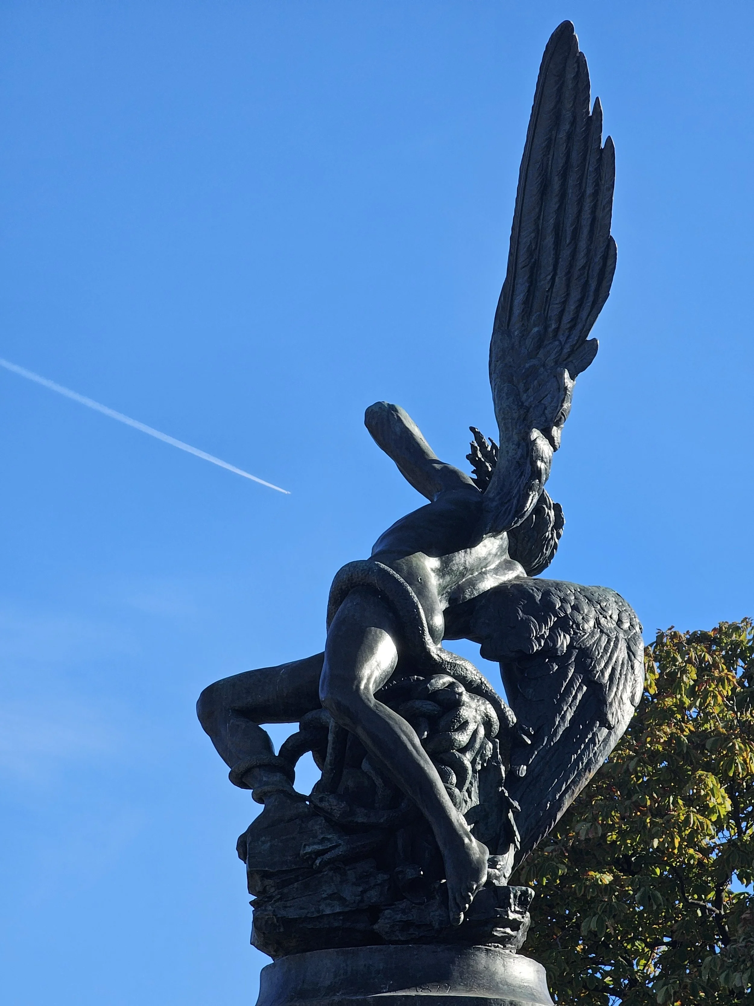 A bronze statue of a mythological figure with large wings, kneeling and holding a snake, set against a clear blue sky with a plane contrail.