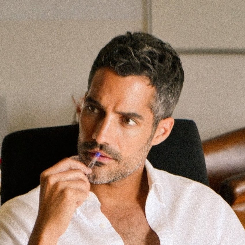 A man with dark, curly hair and beard sitting in a chair, holding a cigarette in his right hand near his lips, wearing a white shirt, looking thoughtful.