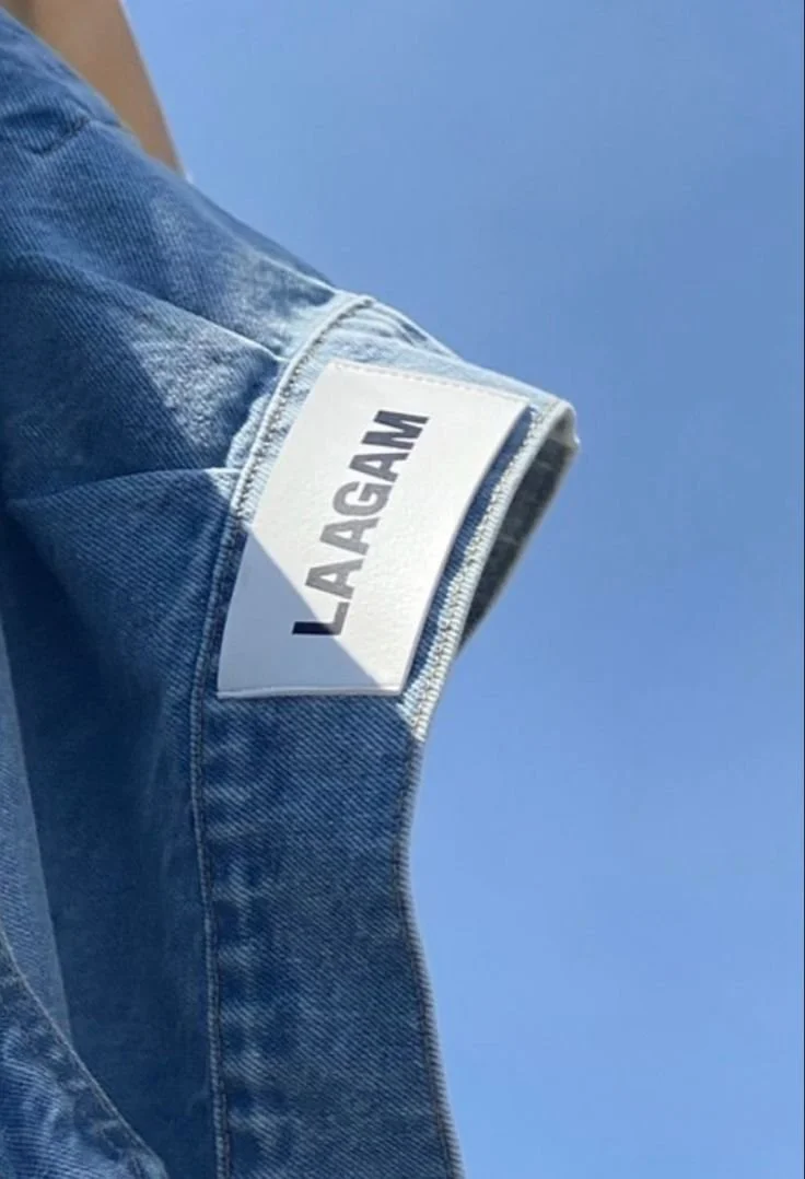 Close-up of a blue denim jacket with a white label on the sleeve that reads "LAAGAM" against a clear blue sky.