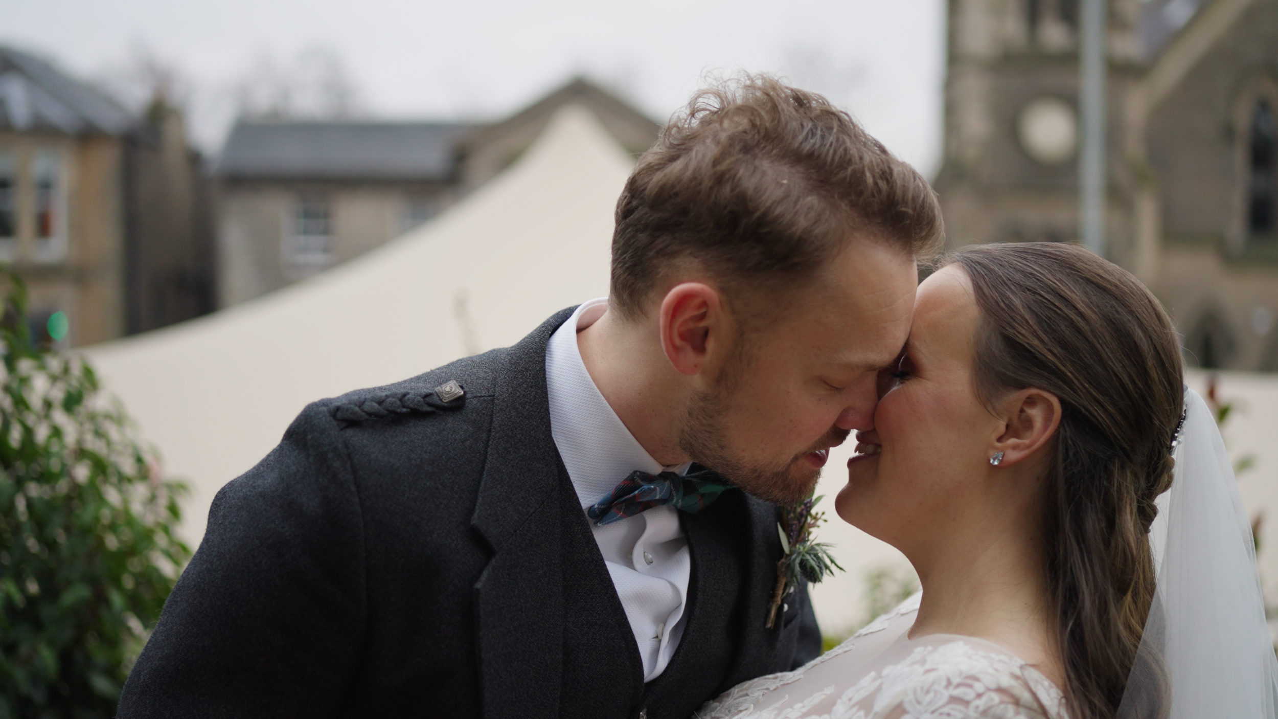 Bride and groom touching foreheads outdoors, smiling, with a blurred church in the background.