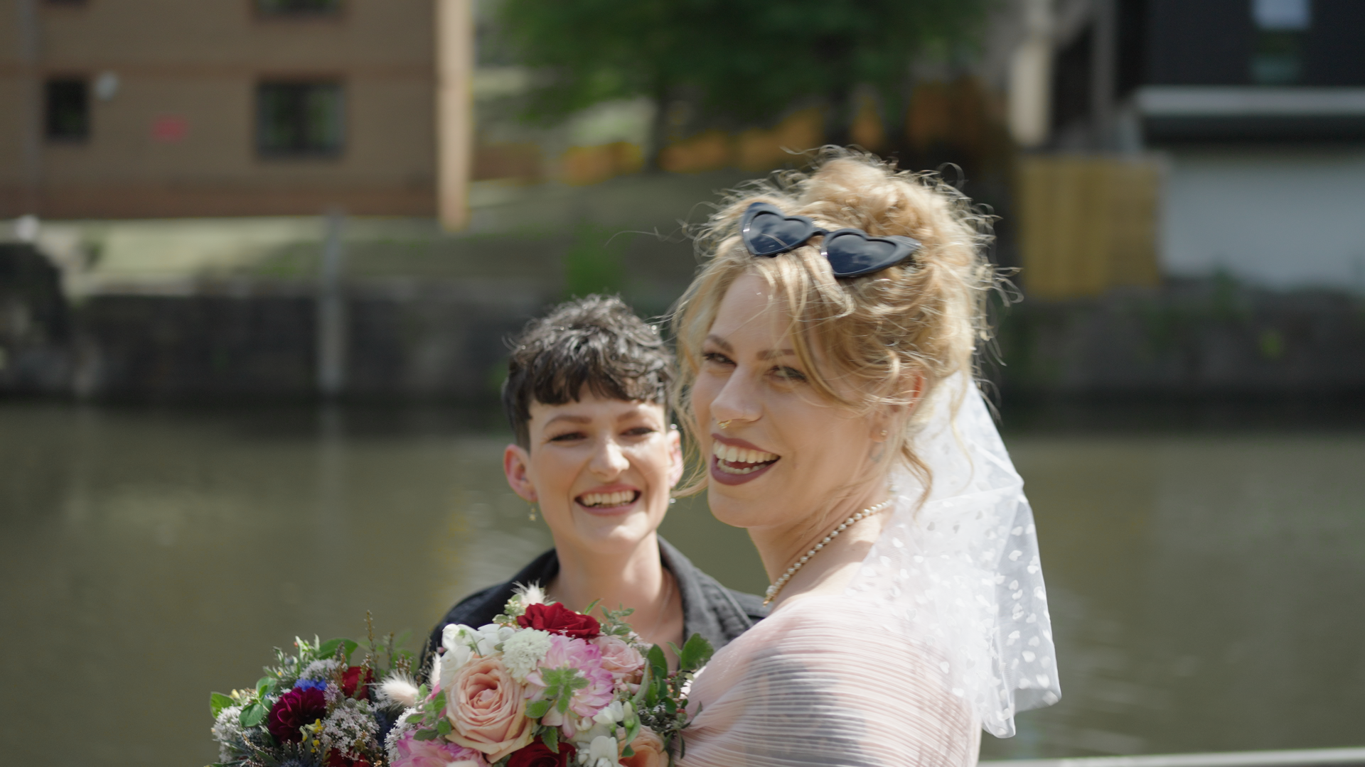 Two women smiling, with one holding a bouquet of flowers. The person on the right is wearing a veil and heart-shaped sunglasses. They are standing outdoors near water.