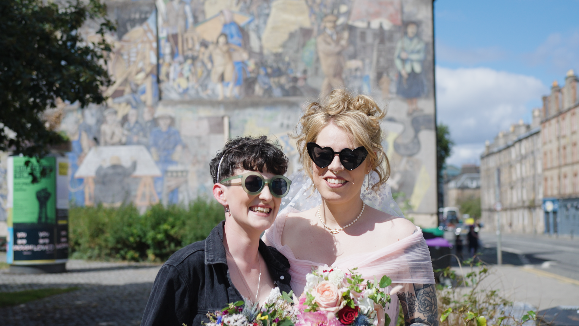 Two women smiling, wearing sunglasses, holding bouquets, standing outdoors in front of a mural.