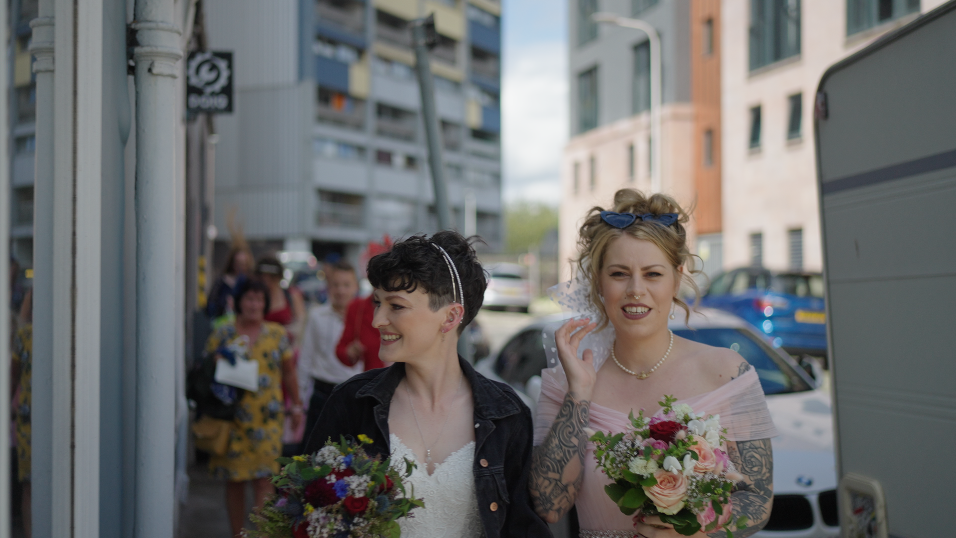 Two women in wedding attire holding bouquets, walking on a street with modern buildings in the background, followed by a group of people.